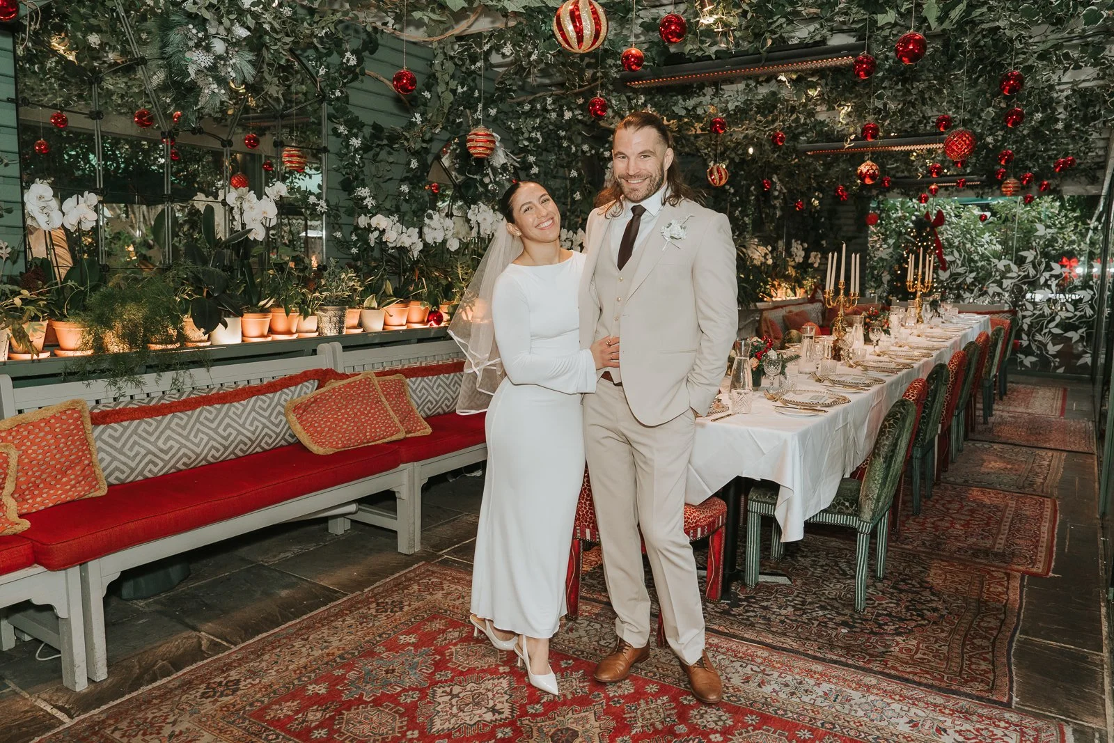  Portrait of the bride and groom standing beside the long dining table in the Glasshouse Room at The Ivy Chelsea Garden during a Christmas wedding celebration 