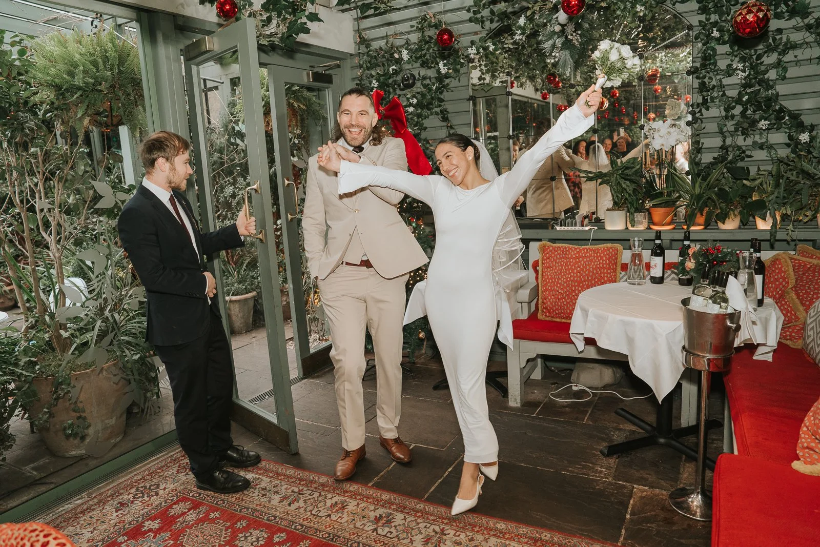  Bride and groom entering their wedding reception in the Glasshouse Room at The Ivy Chelsea Garden, celebrating beneath festive greenery and red Christmas baubles 