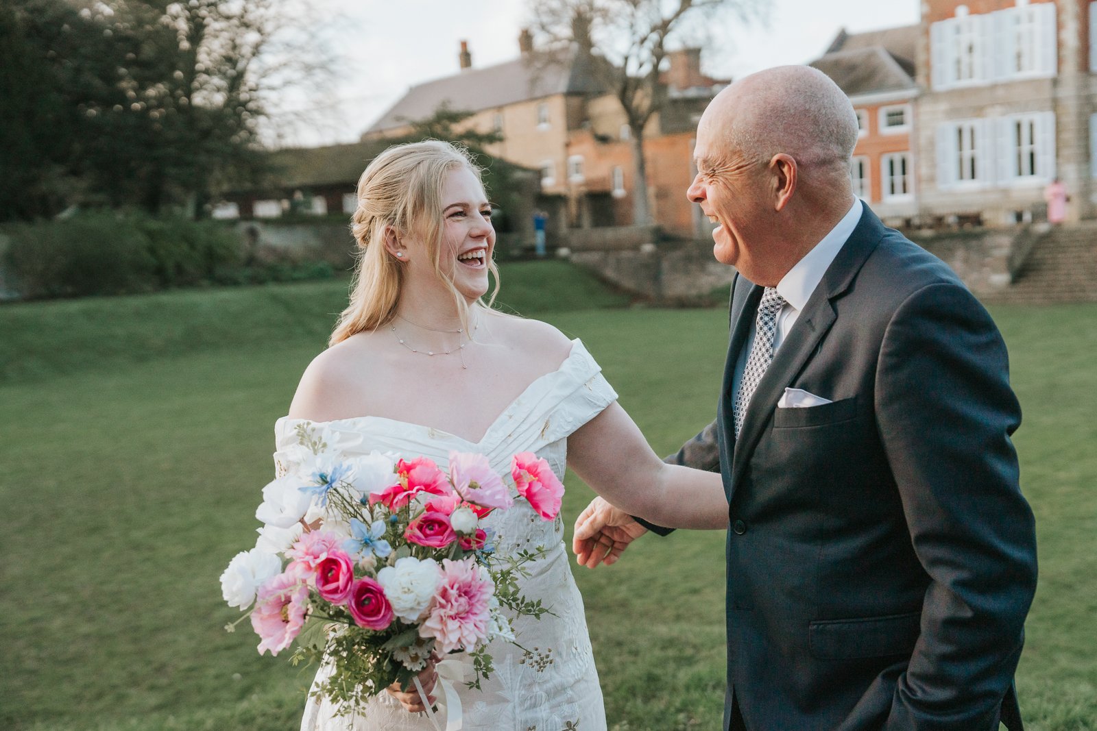  Bride laughing with her dad in the grounds of York House, Twickenham, holding a bright pink and blue bouquet during candid moments after the wedding ceremony. 