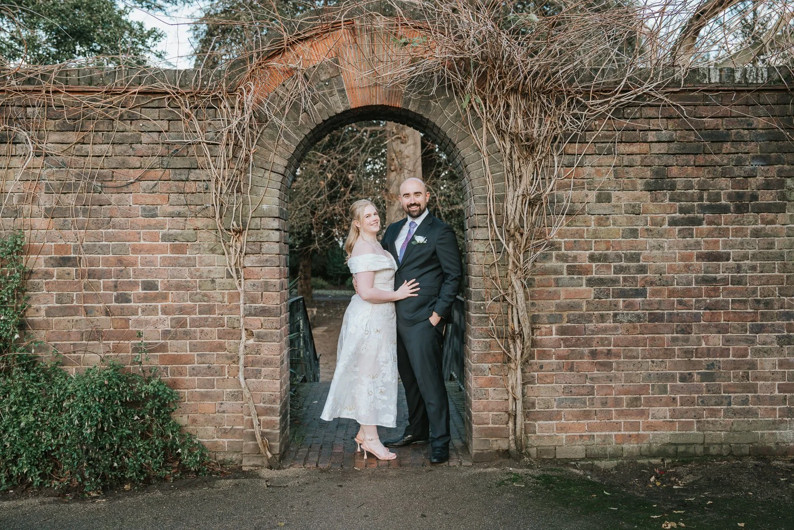  Bride and groom standing together beneath the brick archway in the York House gardens, captured in a classic portrait during their Twickenham wedding photography session. 