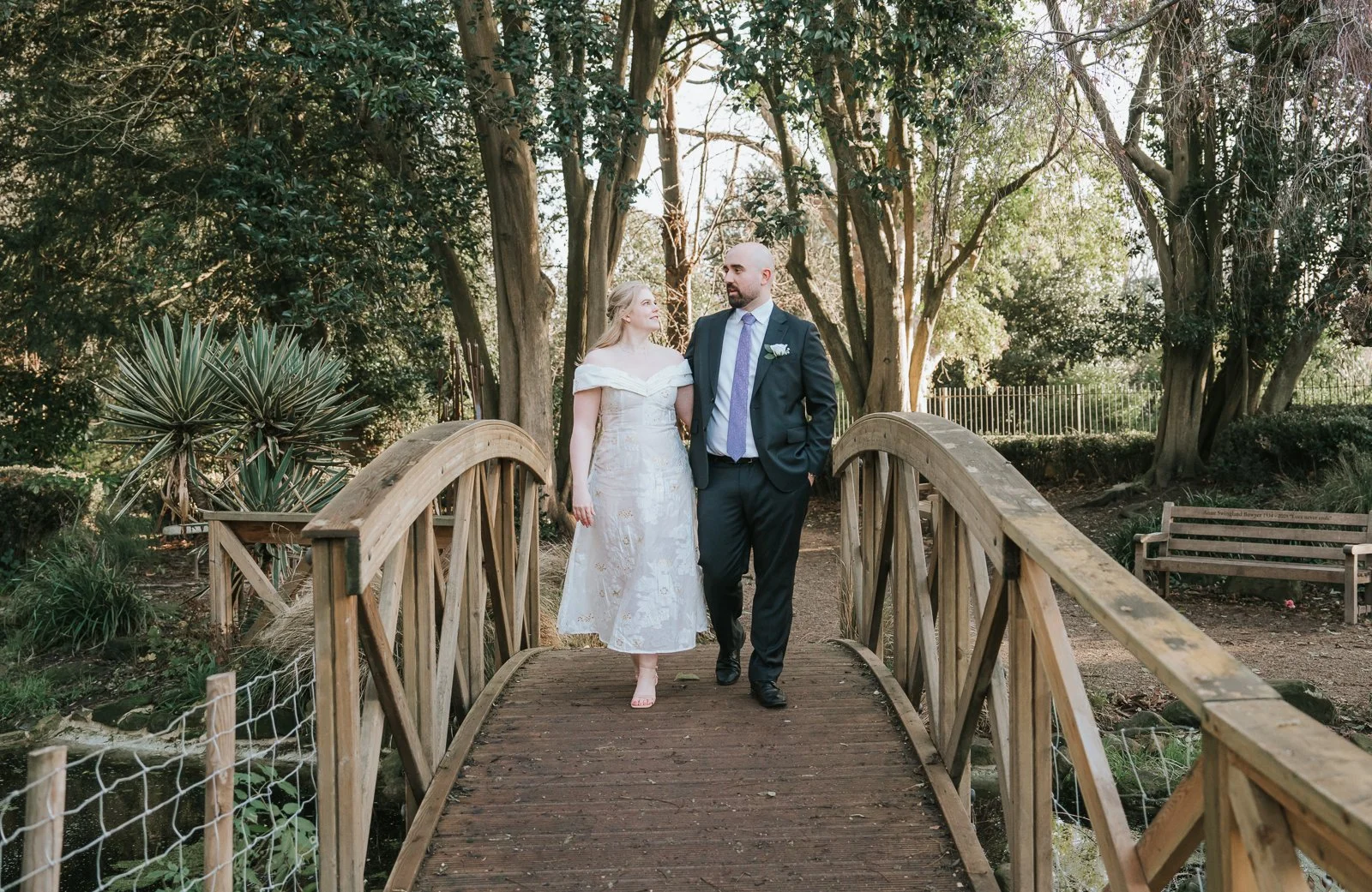  Bride and groom walking arm in arm across the wooden bridge in the gardens at York House, Twickenham, during relaxed outdoor wedding portraits after their register office ceremony. 