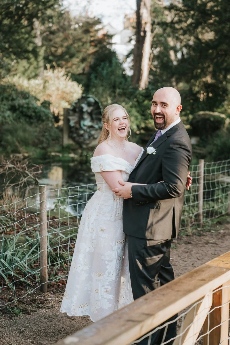  Joyful bride and groom laughing together in the York House gardens in Twickenham, captured in a candid moment during their natural, documentary-style wedding photography. 