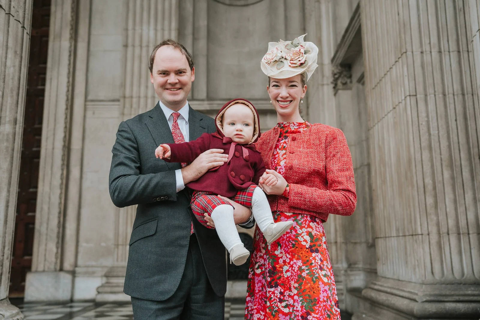  Family portrait outside St Paul’s Cathedral after a christening, parents holding their baby in front of the grand stone columns 
