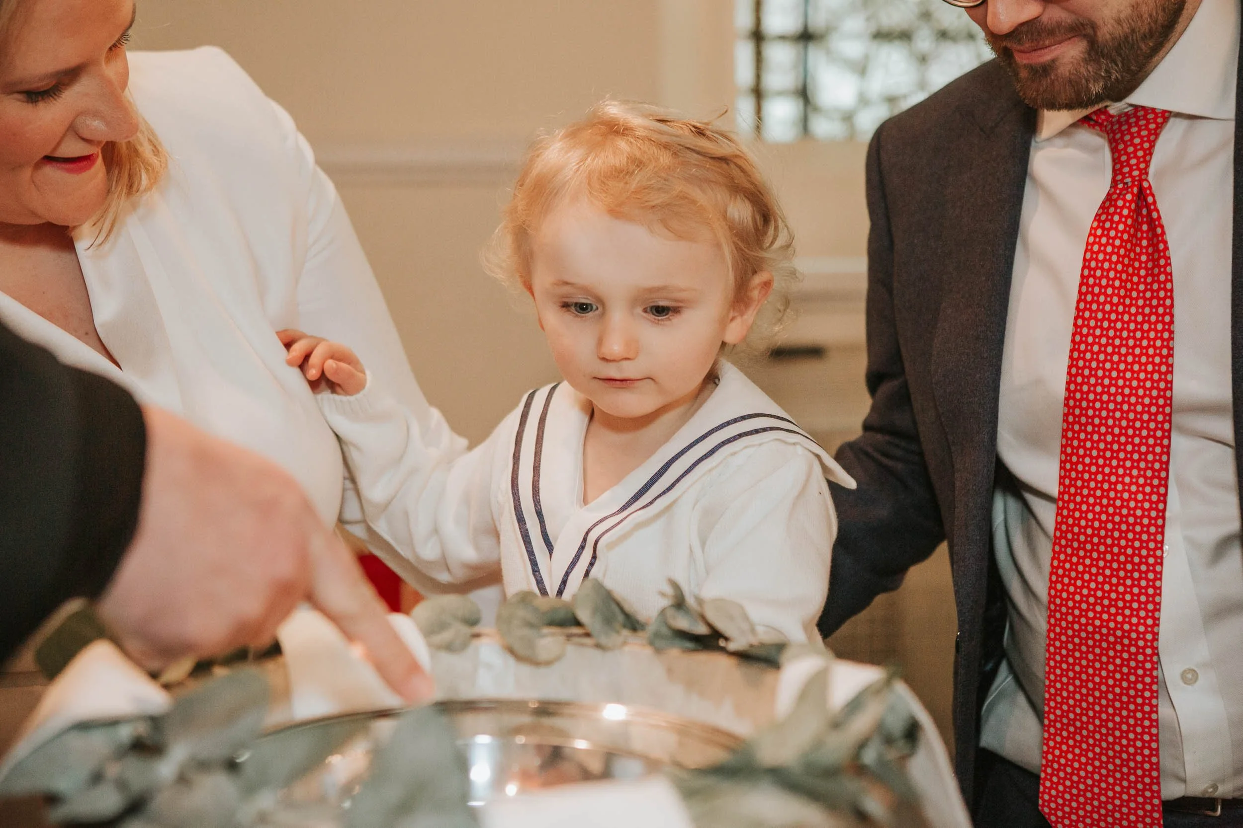  Toddler watching baptism ceremony at christening reception with parents gathered around in London. 
