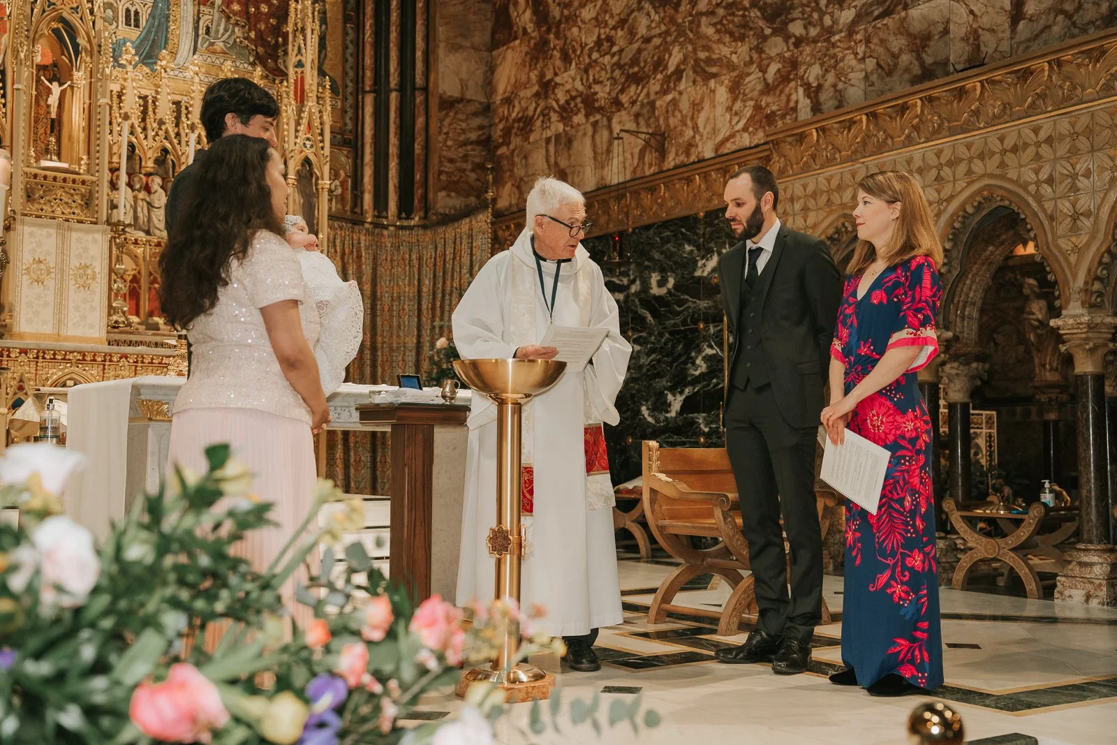  Priest speaking to parents and godparents during Catholic christening ceremony inside ornate London church. 
