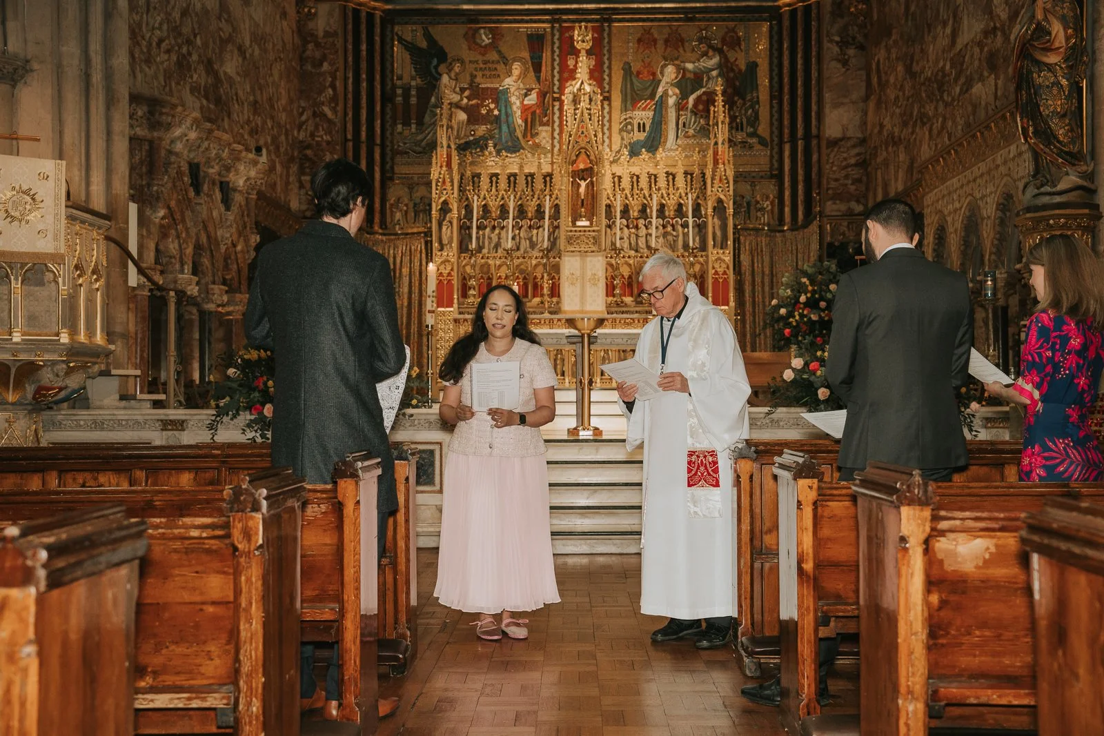  Parents and godparents standing during prayers at Catholic baptism ceremony inside London church with decorative altar behind. 