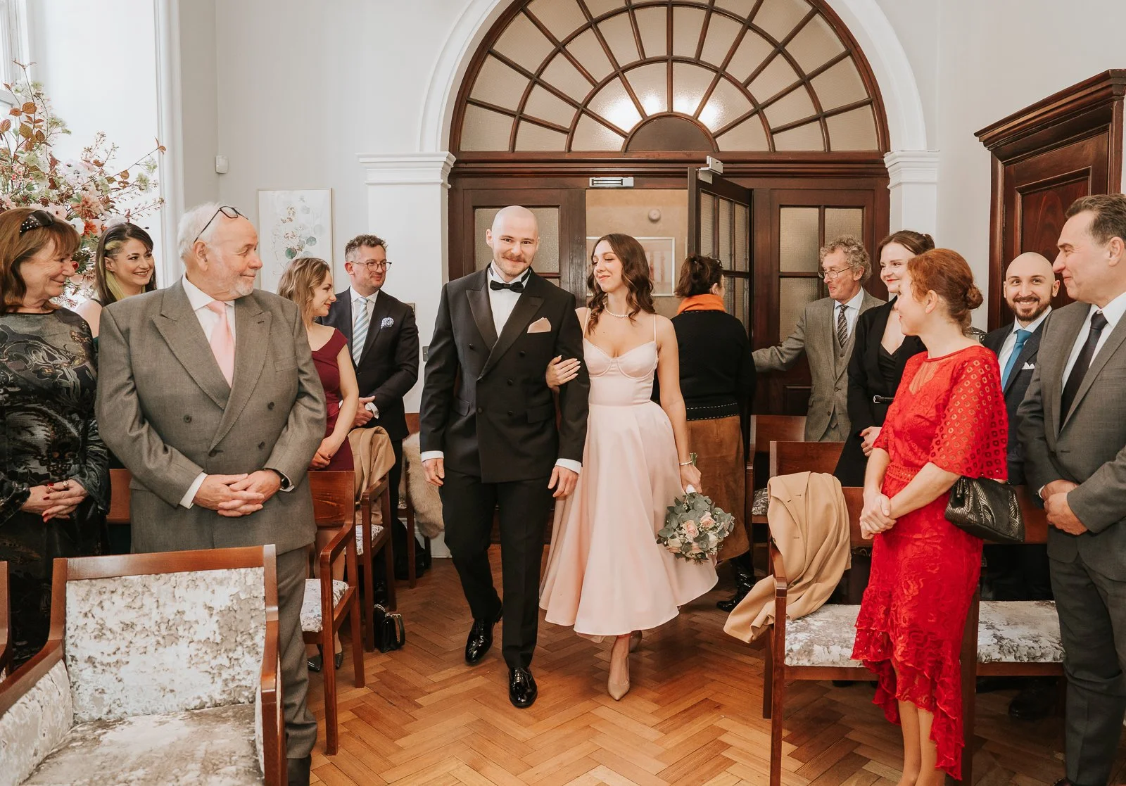  Bride and groom walking down the aisle after their Rossetti Room ceremony at Chelsea Old Town Hall, surrounded by smiling wedding guests. 