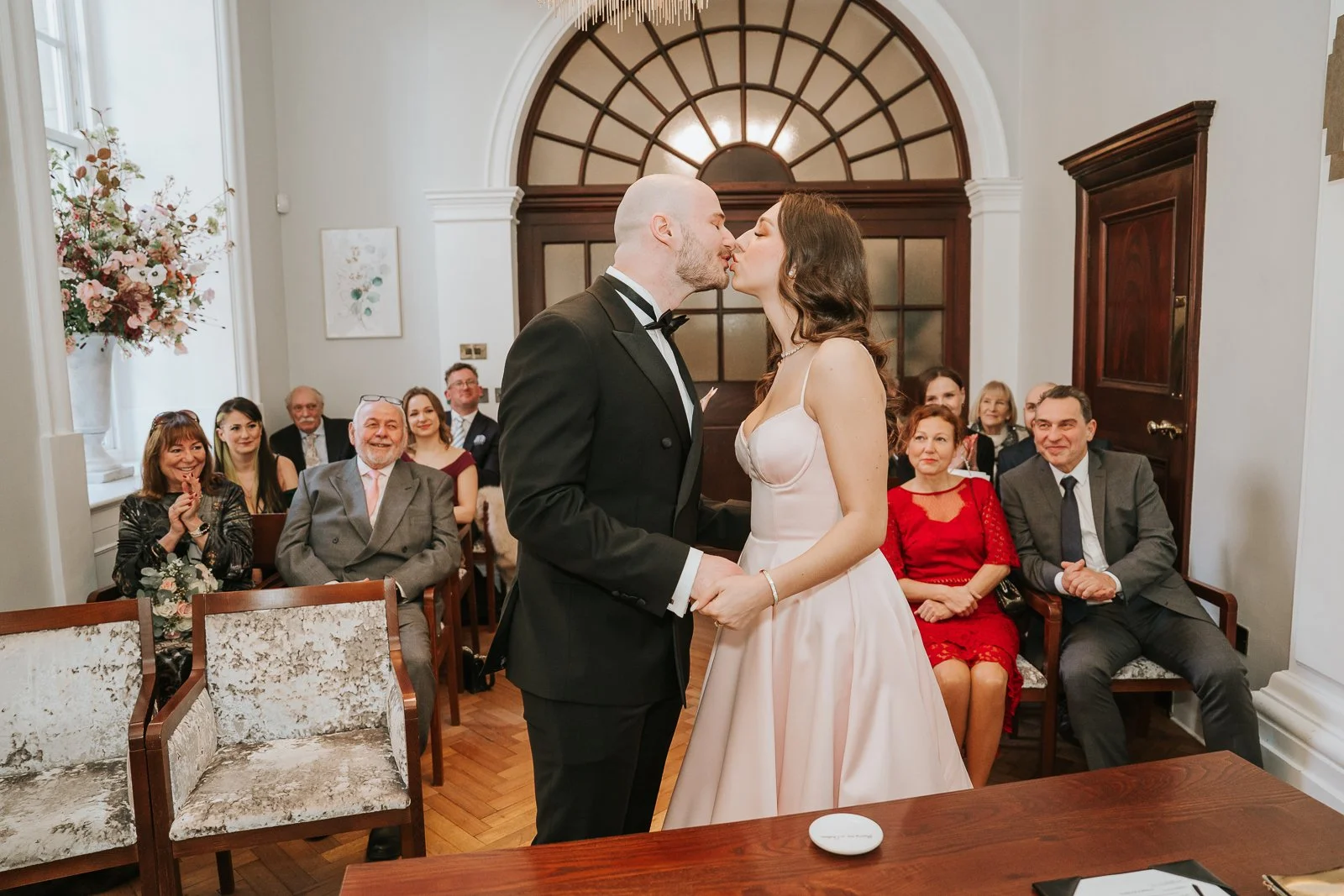  First kiss as newlyweds inside the Rossetti Room at Chelsea Old Town Hall with guests applauding behind them. 