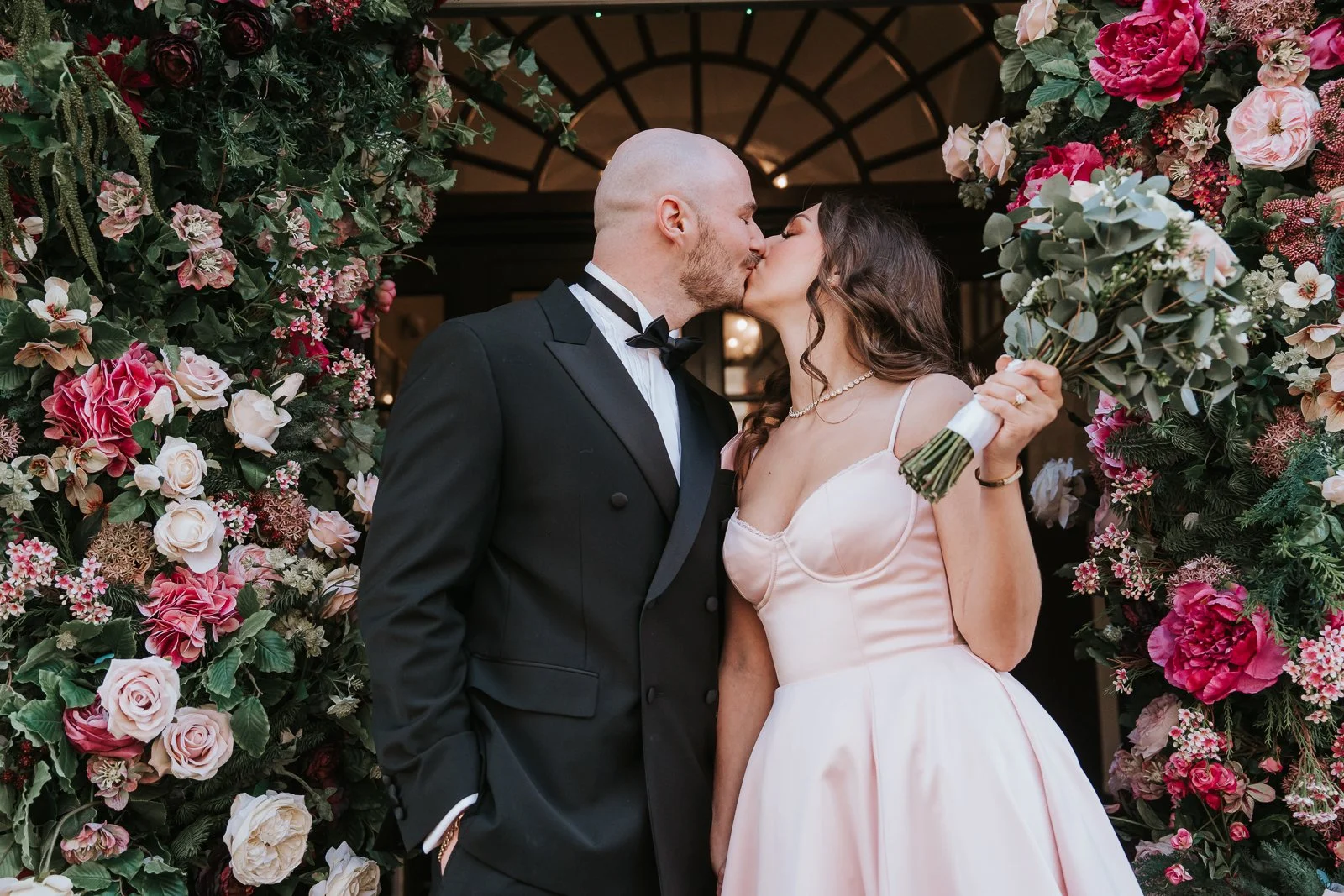  Newlyweds kissing outside after their intimate civil wedding ceremony with floral entrance backdrop. 