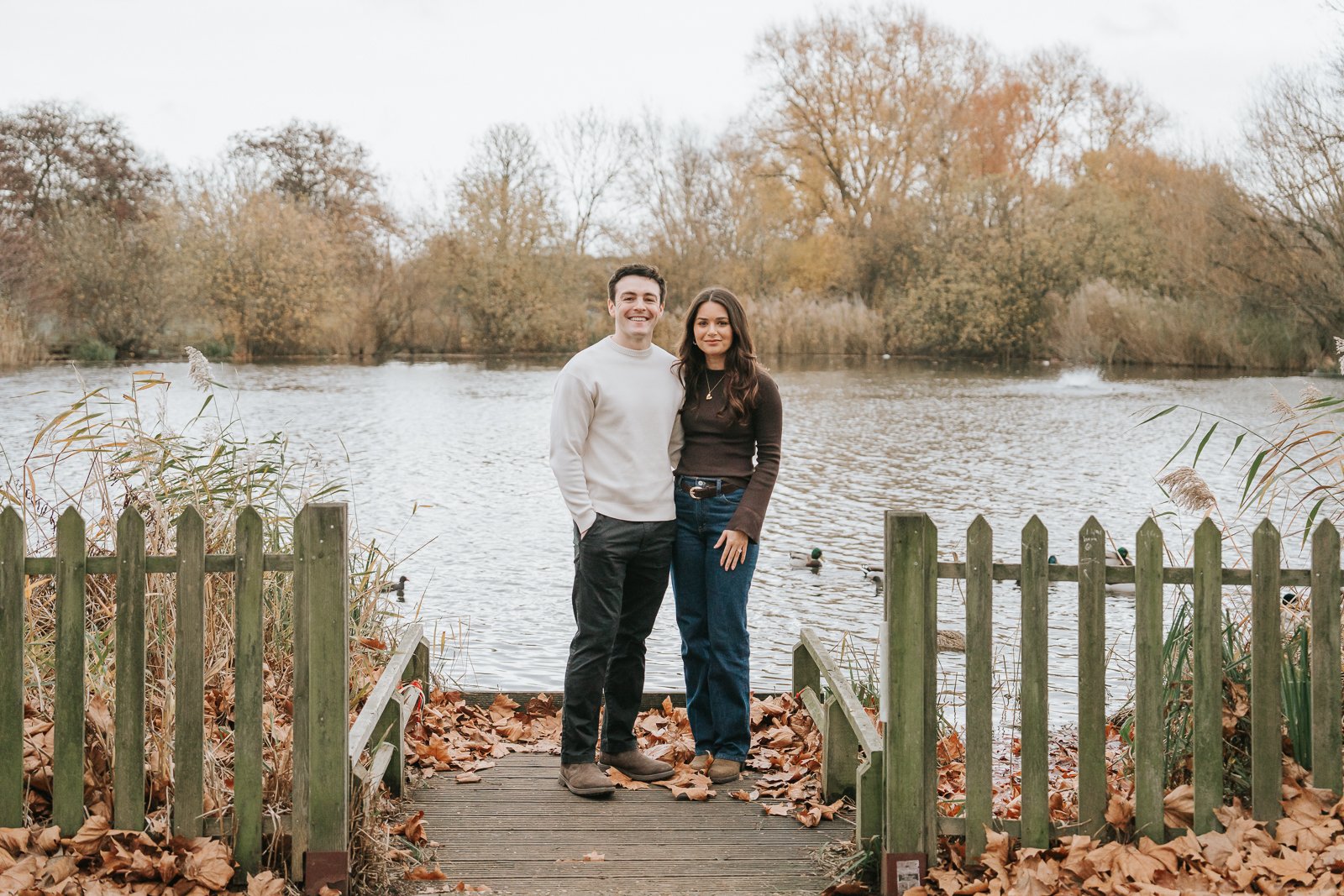  Newly engaged couple standing on wooden jetty overlooking Clapham Common pond in autumn, South London proposal setting. 