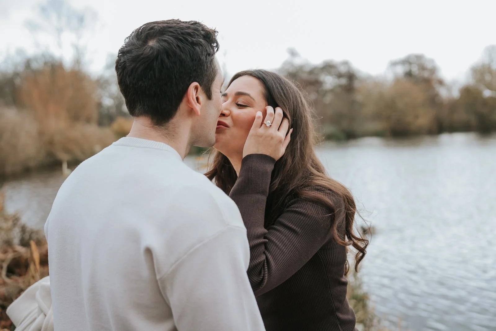  Couple sharing a kiss by the pond at Clapham Common after a romantic London proposal, autumn trees reflected in the water. 