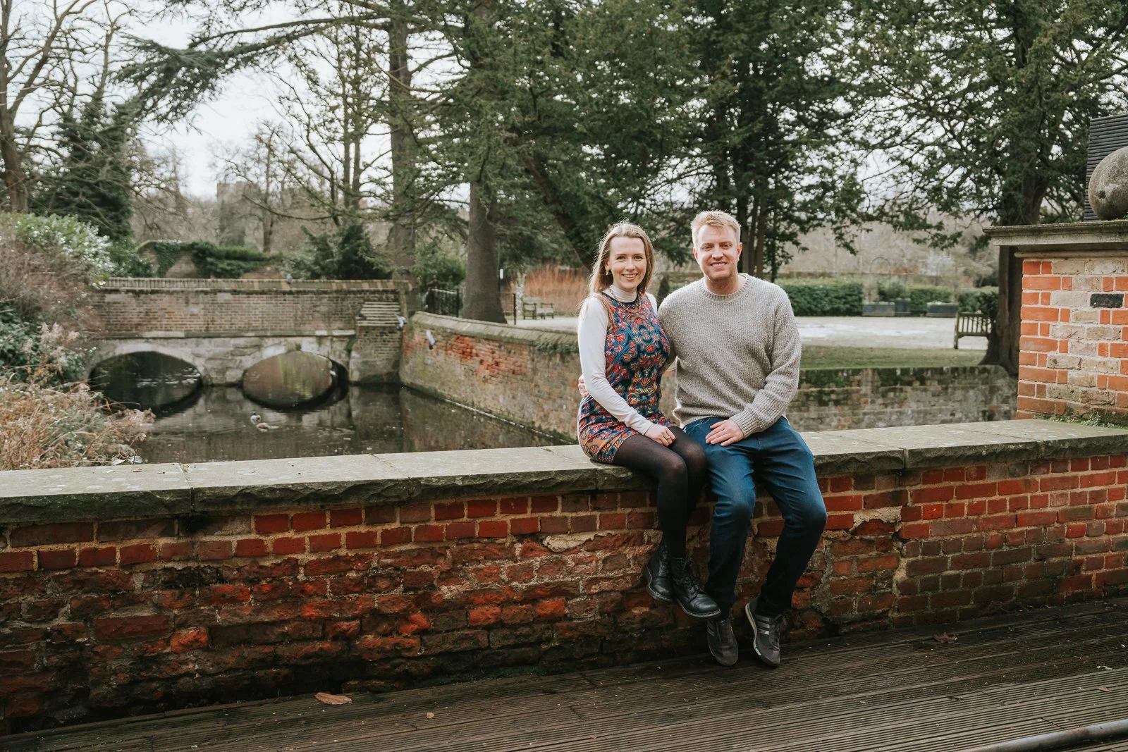  Engaged couple sitting on a red brick wall overlooking the moat and gardens at Eltham Tudor Barn, relaxed South East London engagement photo session. 