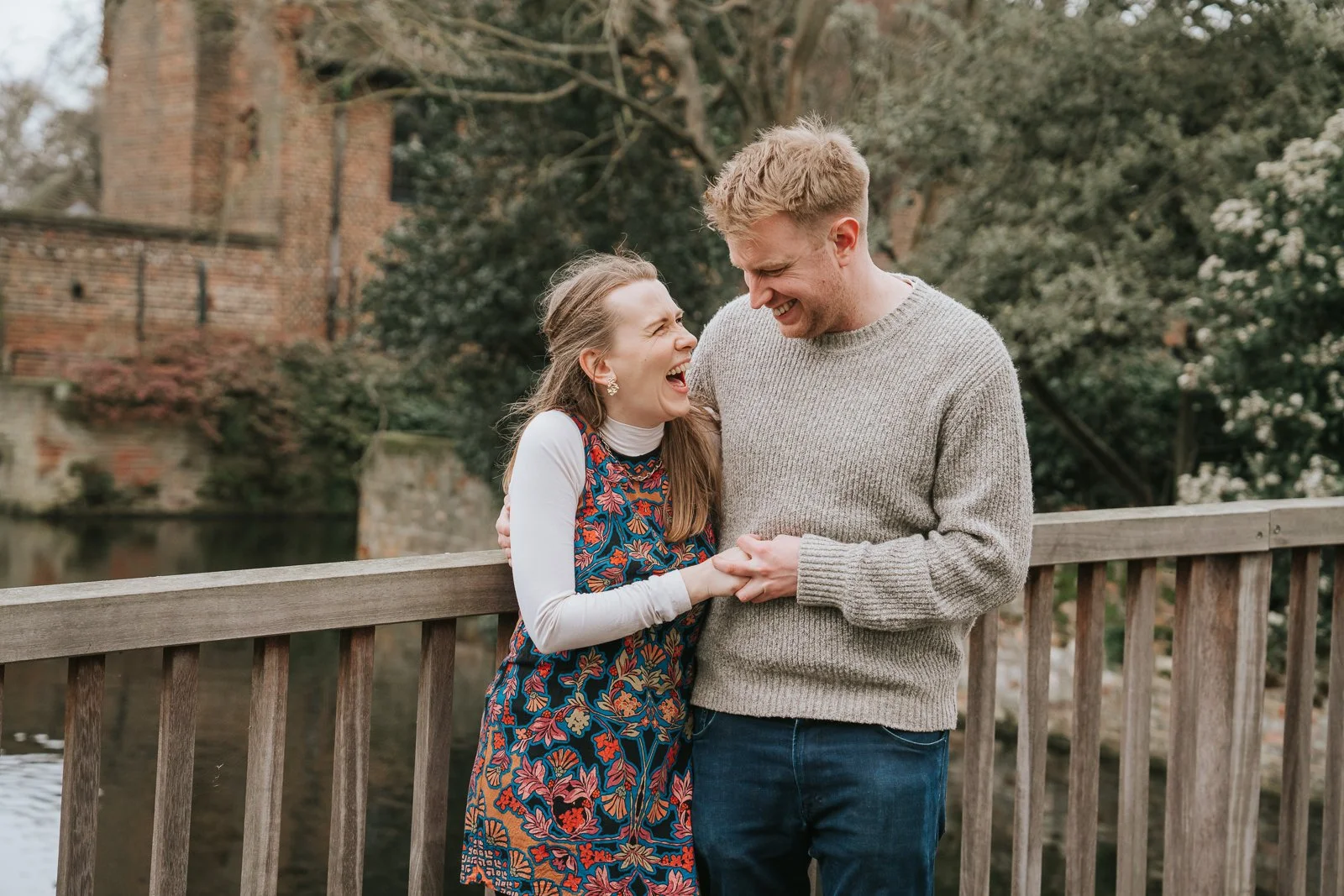  Couple laughing together on the wooden bridge over the moat at Eltham Tudor Barn in South East London, relaxed engagement photo with historic brick backdrop. 