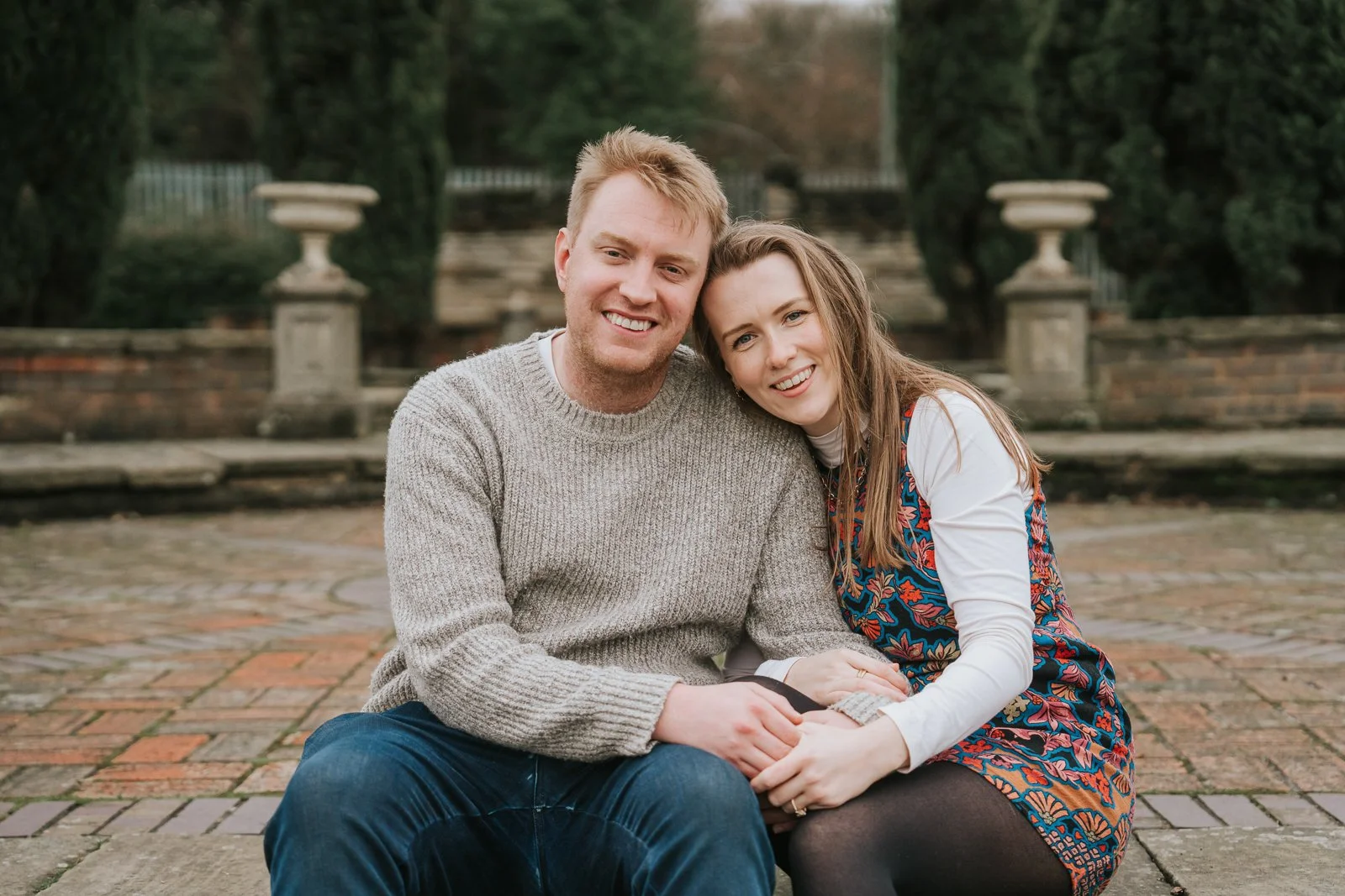  Couple sitting close together in the formal gardens at Eltham Tudor Barn, romantic engagement portrait at a historic London wedding venue. 