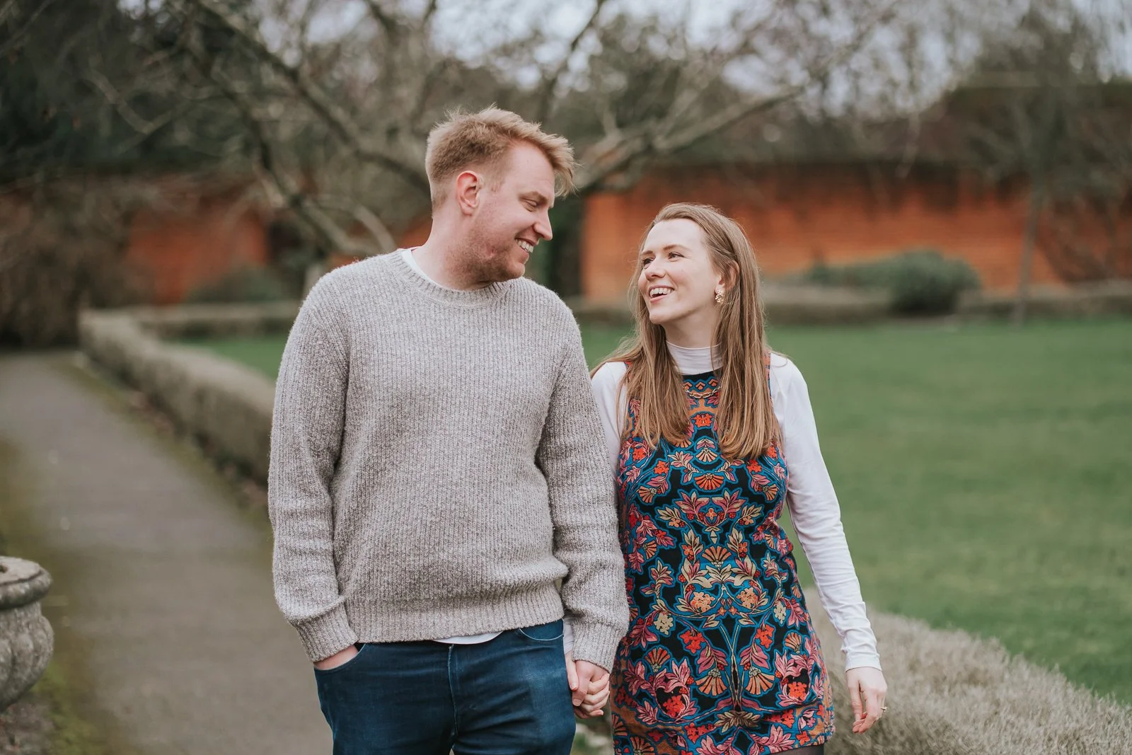  Engaged couple strolling through the landscaped grounds of Eltham Tudor Barn, South East London pre-wedding photography with historic red brick walls in the background. 