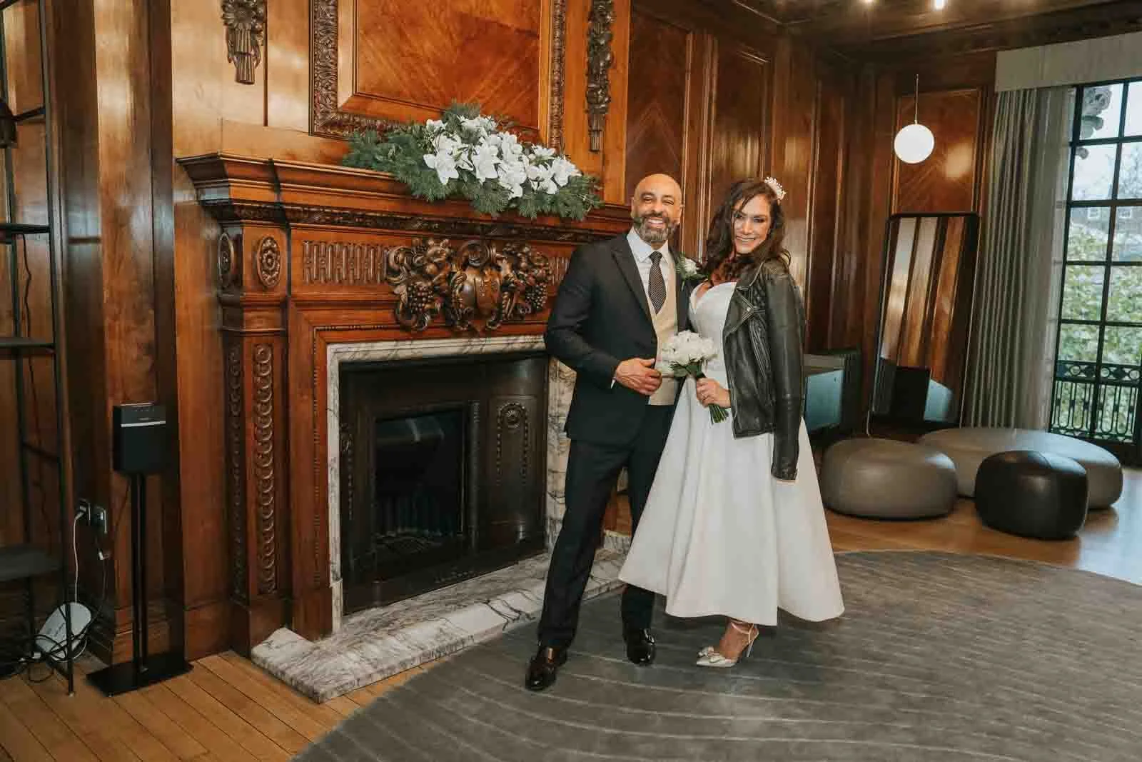  Bride and groom posing beside the ornate fireplace inside Old Marylebone Town Hall in London, with wood-panelled walls and winter florals, intimate London registry office wedding. 