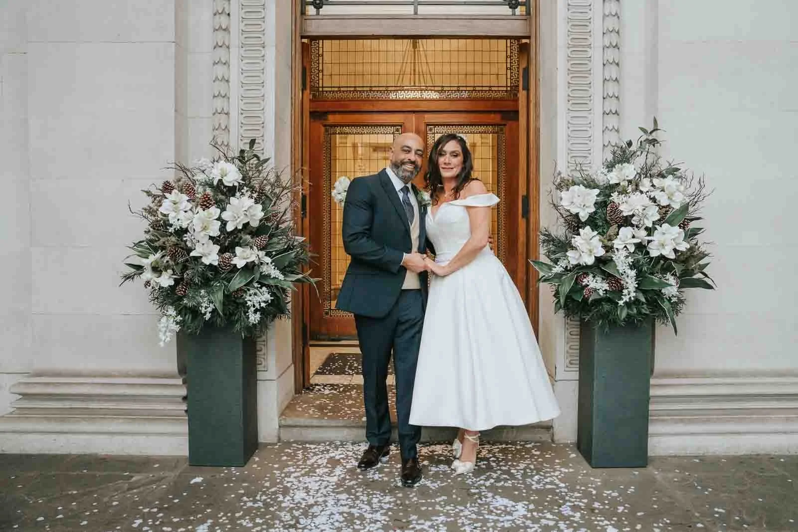  Bride and groom standing between large floral arrangements at the entrance of Old Marylebone Town Hall, romantic London civil ceremony confetti moment. 
