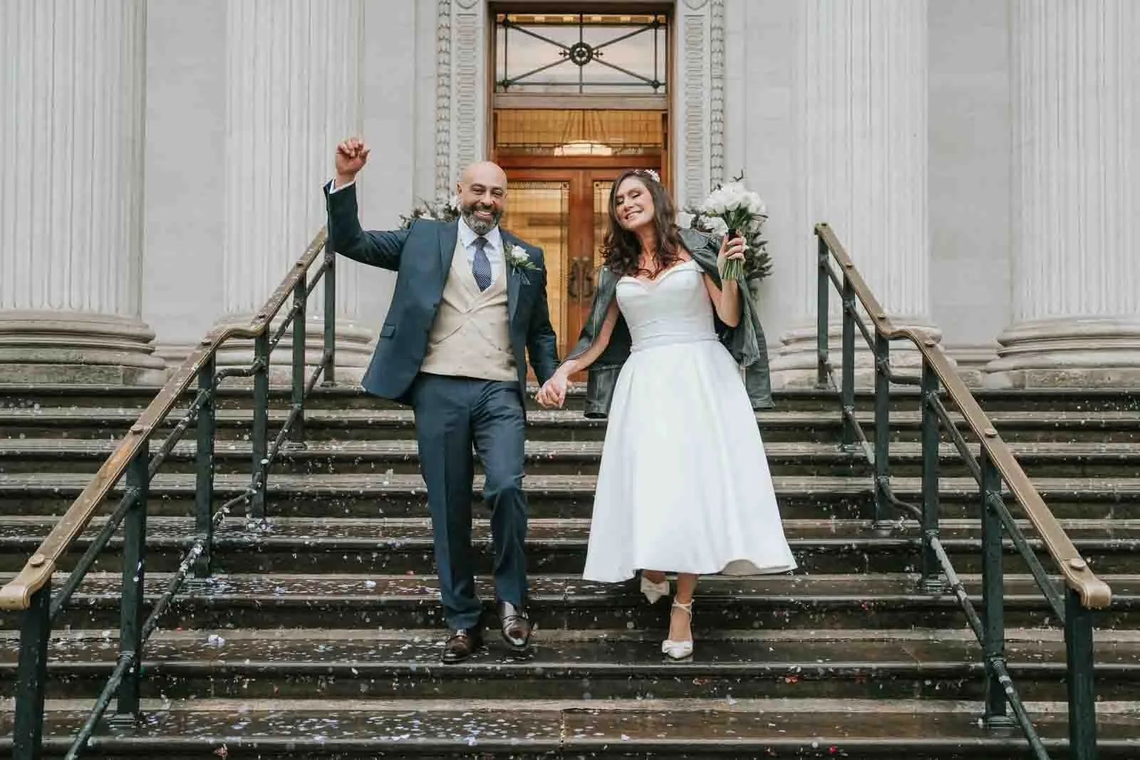  Newly married couple walking down the steps of Old Marylebone Town Hall, holding hands and celebrating with confetti, iconic London registry office wedding photo. 
