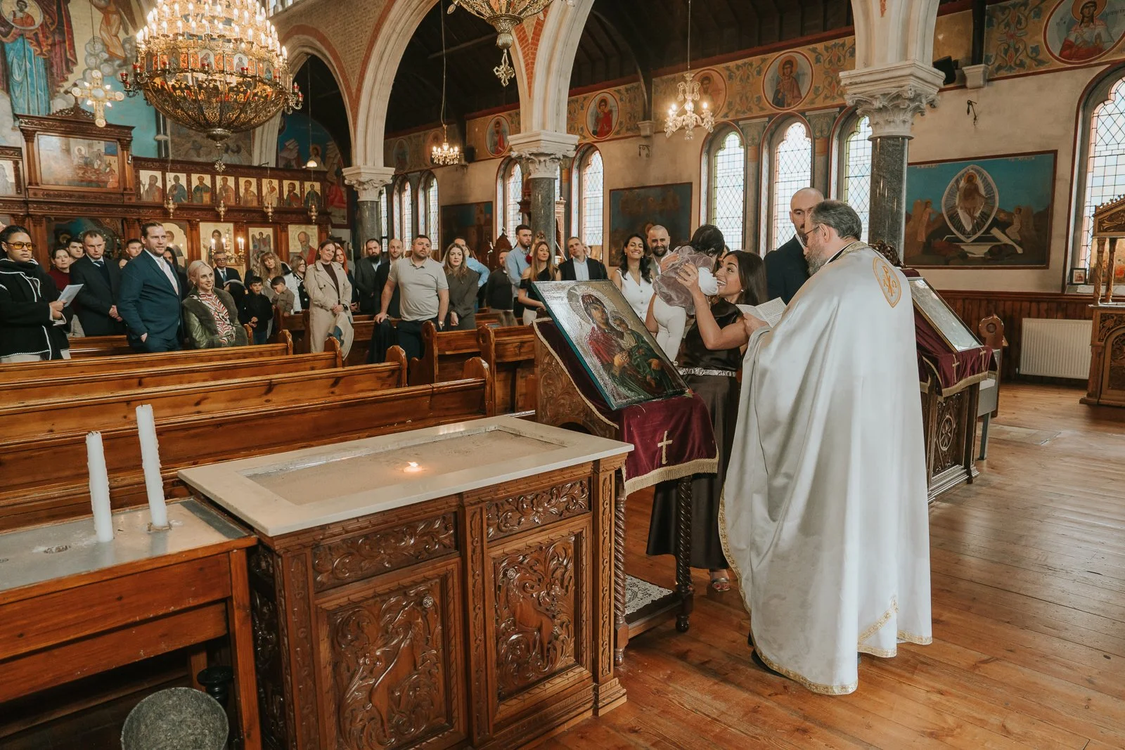  Greek Orthodox christening ceremony at St Constantine &amp; St Helen Church in Crystal Palace, with priest blessing baby at the font surrounded by family and guests inside the icon-filled South London church. 