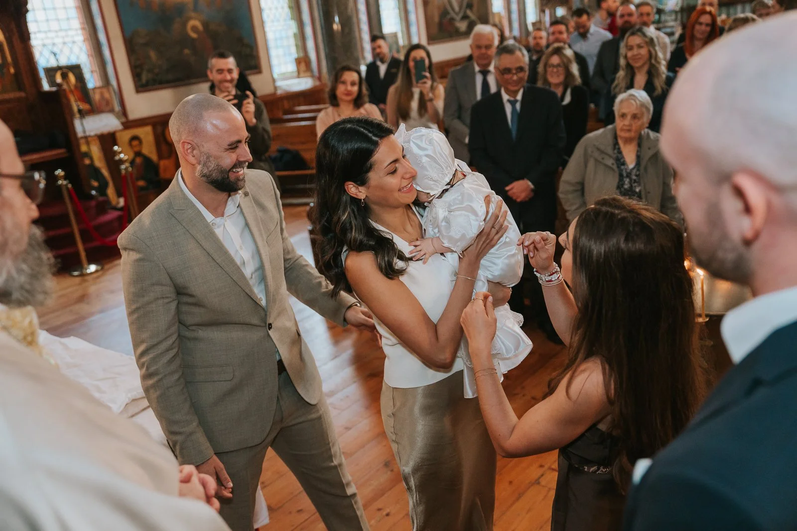  Mother holding her baby in a traditional christening gown as guests look on inside St Constantine &amp; St Helen Church in SE19. 