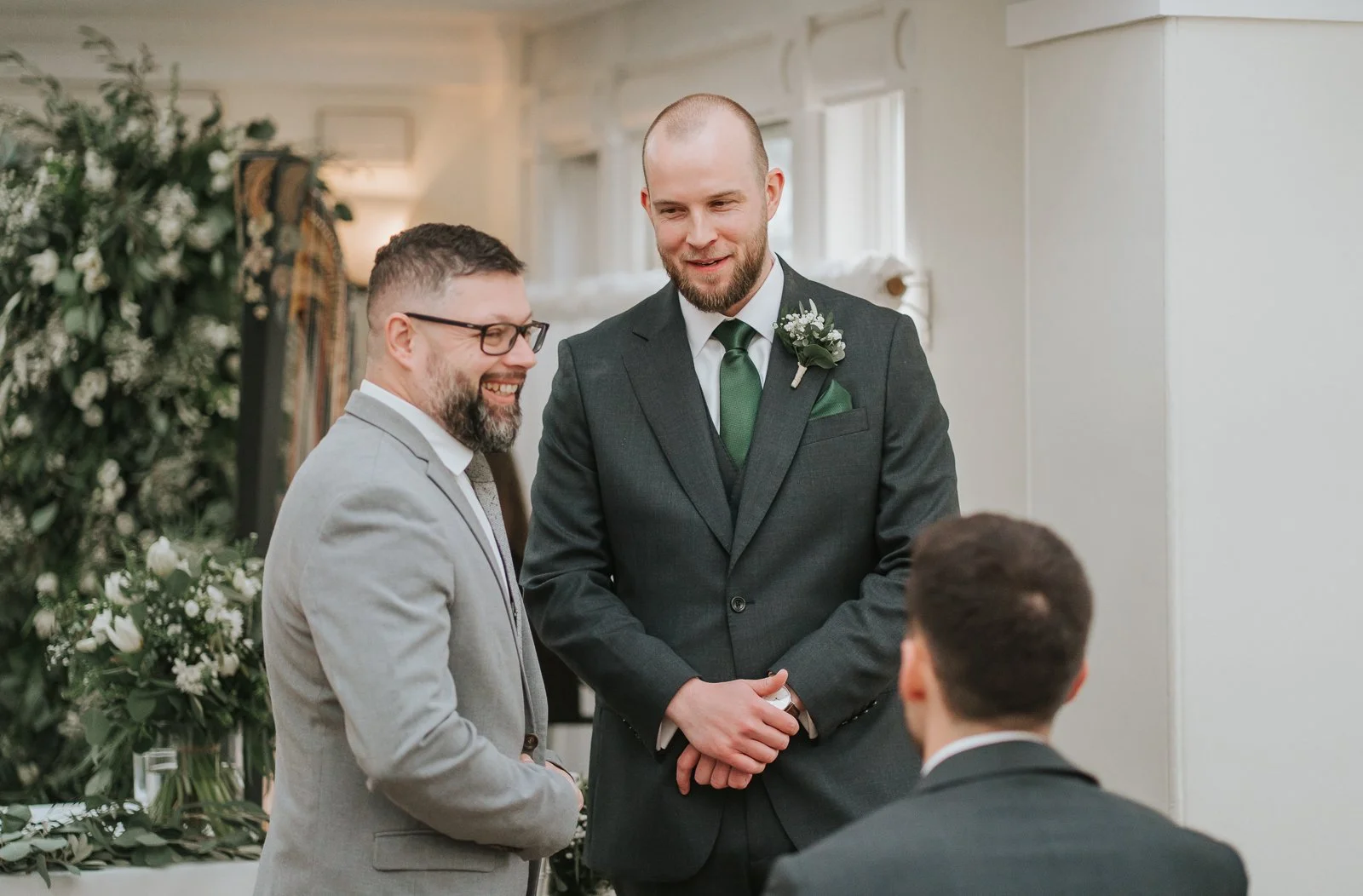  Groom chatting with his best man before the ceremony inside the Belvedere Suite at Pembroke Lodge, Richmond Park wedding venue with natural light and greenery décor. 