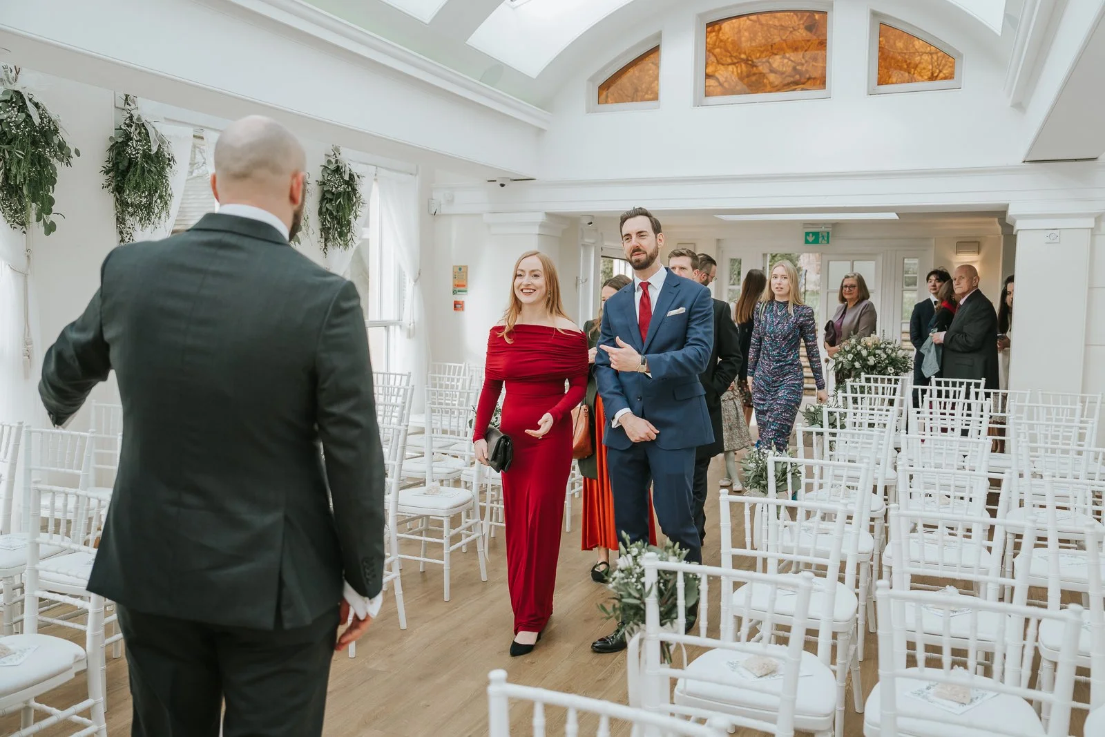  Wedding guests entering the Belvedere Suite at Pembroke Lodge in Richmond Park before a civil ceremony, bright conservatory-style ceremony room with white chiavari chairs. 