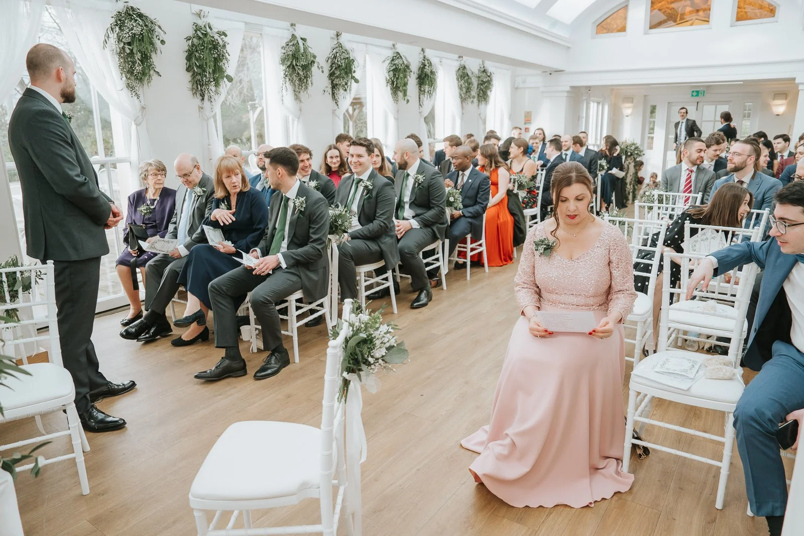  Wedding guests seated inside the Belvedere Suite at Pembroke Lodge awaiting the ceremony, bright Richmond Park wedding venue with floral aisle décor. 