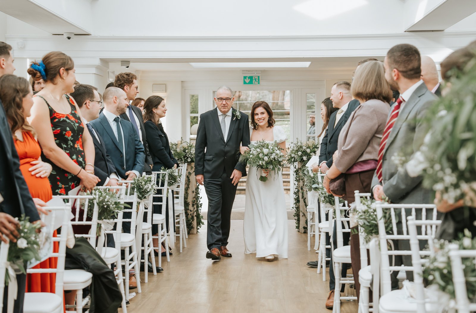  Bride walking down the aisle with her father in the Belvedere Suite at Pembroke Lodge, Richmond Park civil wedding ceremony surrounded by family and friends. 
