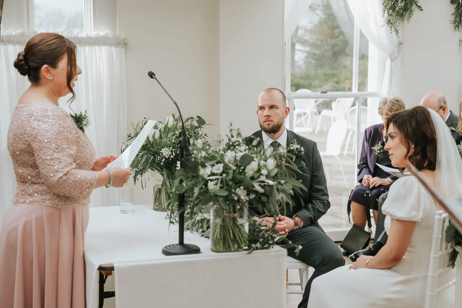  Wedding reading taking place during a Pembroke Lodge ceremony in the Belvedere Suite, Richmond Park wedding venue with greenery and white décor. 