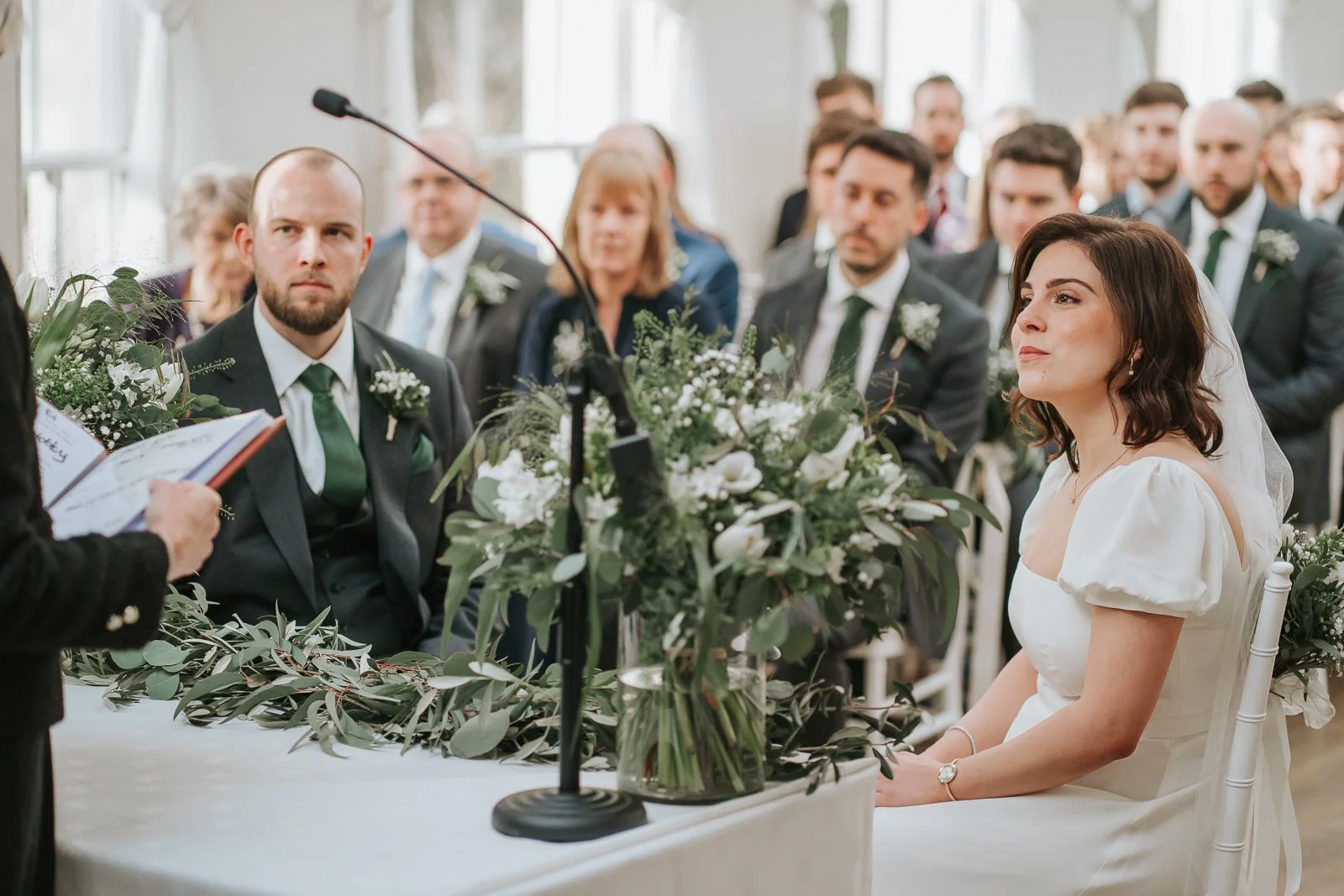  Bride and groom listening to a ceremony reading inside the Belvedere Suite at Pembroke Lodge in Richmond Park, intimate London civil wedding moment. 