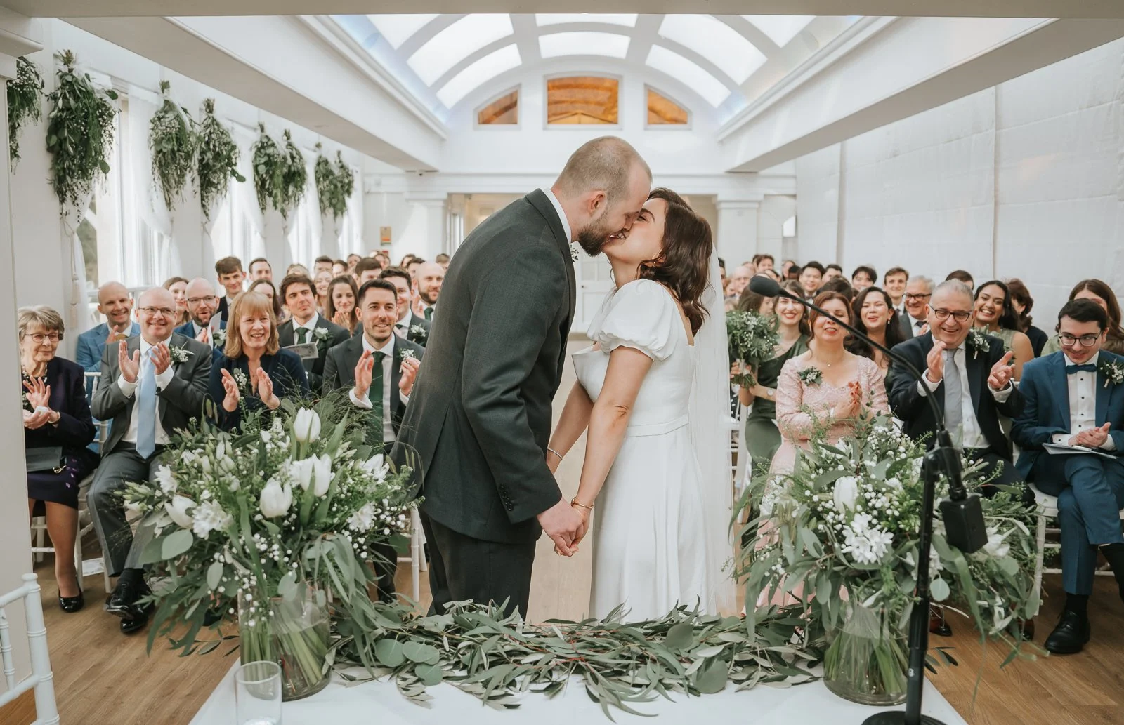  Bride and groom sharing their first kiss during a civil ceremony in the Belvedere Suite at Pembroke Lodge, Richmond Park wedding venue surrounded by guests. 