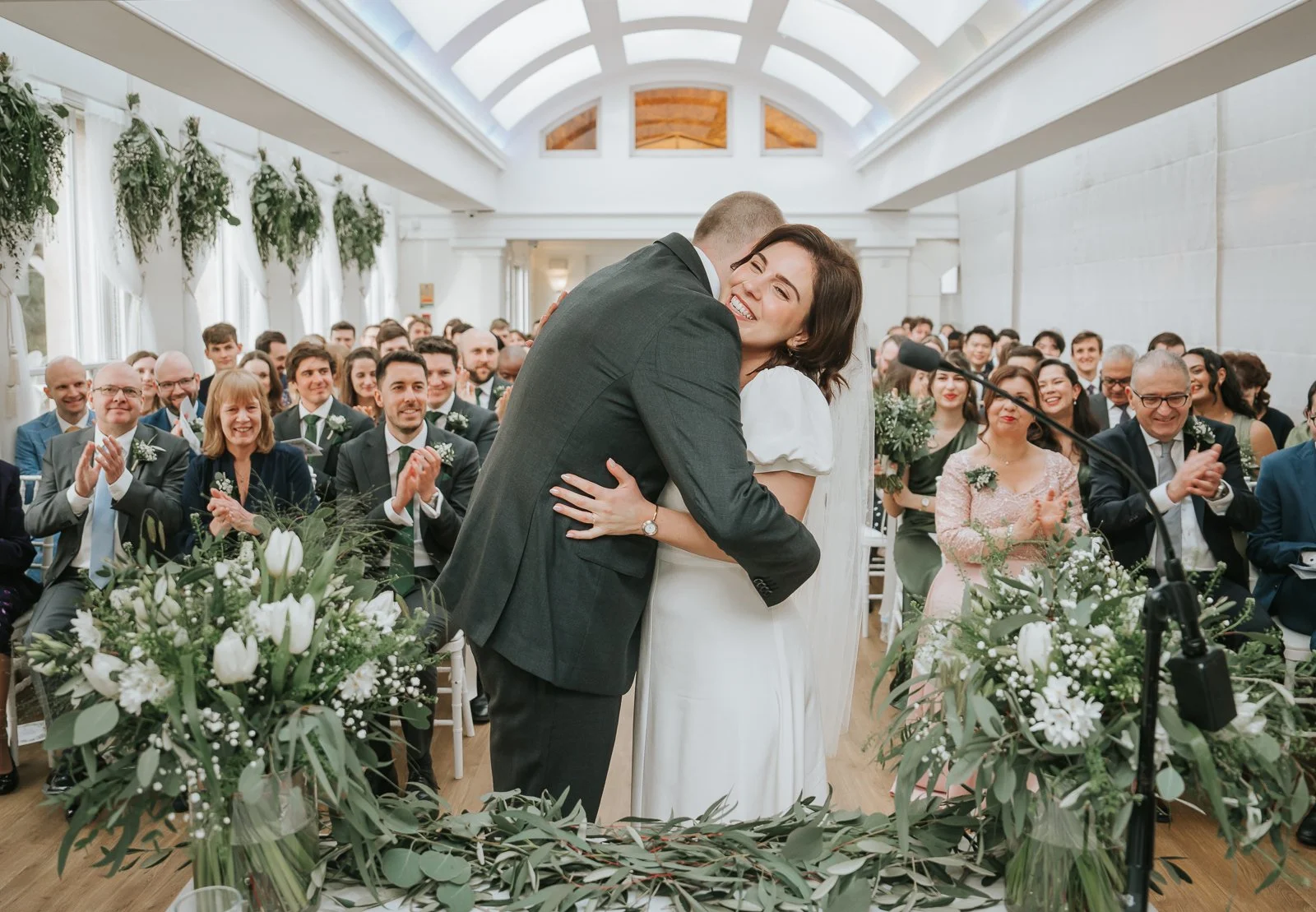  Newly married couple embracing after their ceremony at Pembroke Lodge in the Belvedere Suite, bright Richmond Park wedding venue filled with family and friends. 