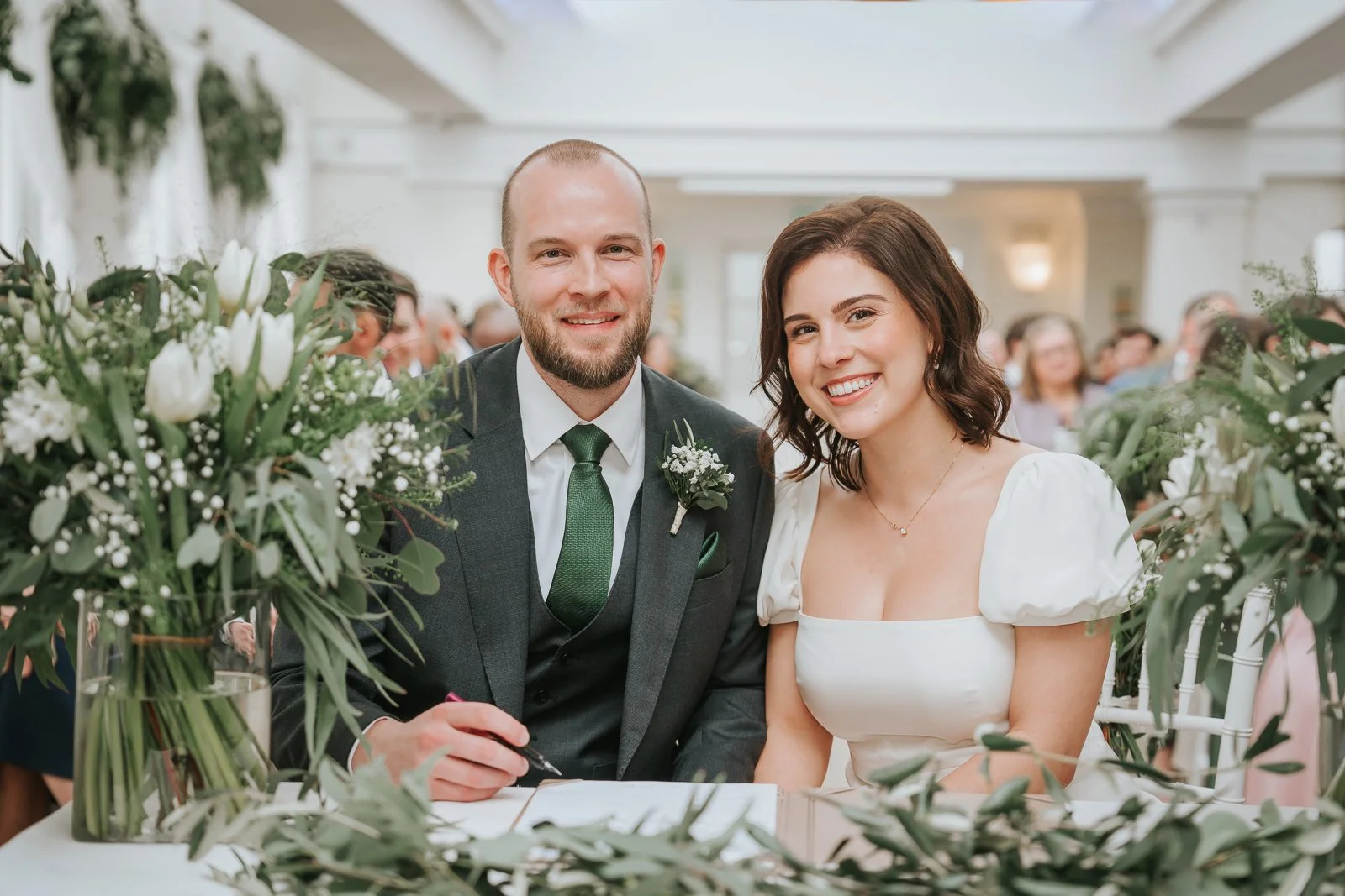  Bride and groom signing the wedding register inside the Belvedere Suite at Pembroke Lodge, Richmond Park wedding ceremony room decorated with greenery. 