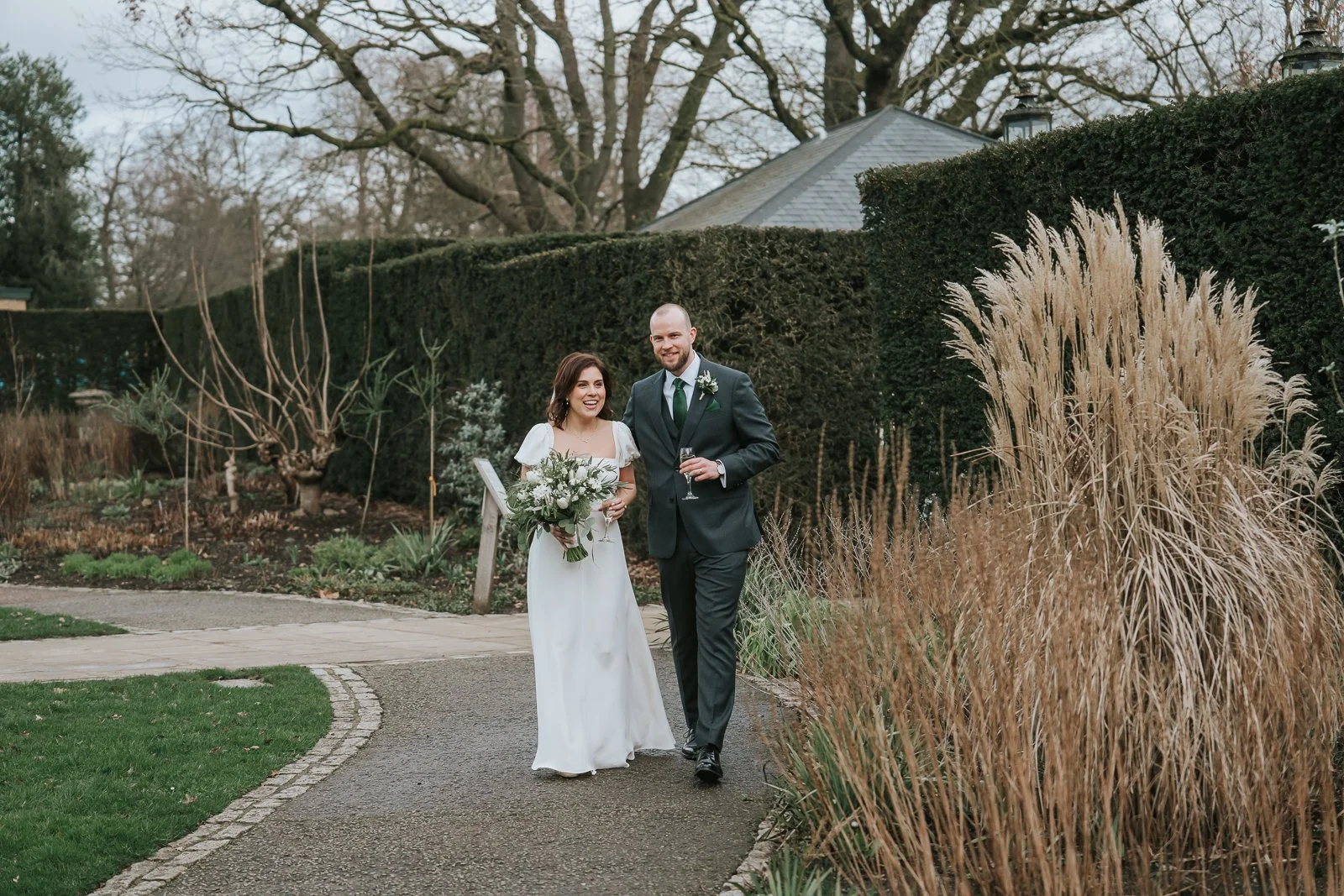  Bride and groom walking through the gardens at Pembroke Lodge in Richmond Park after their wedding ceremony, relaxed South West London wedding photography. 