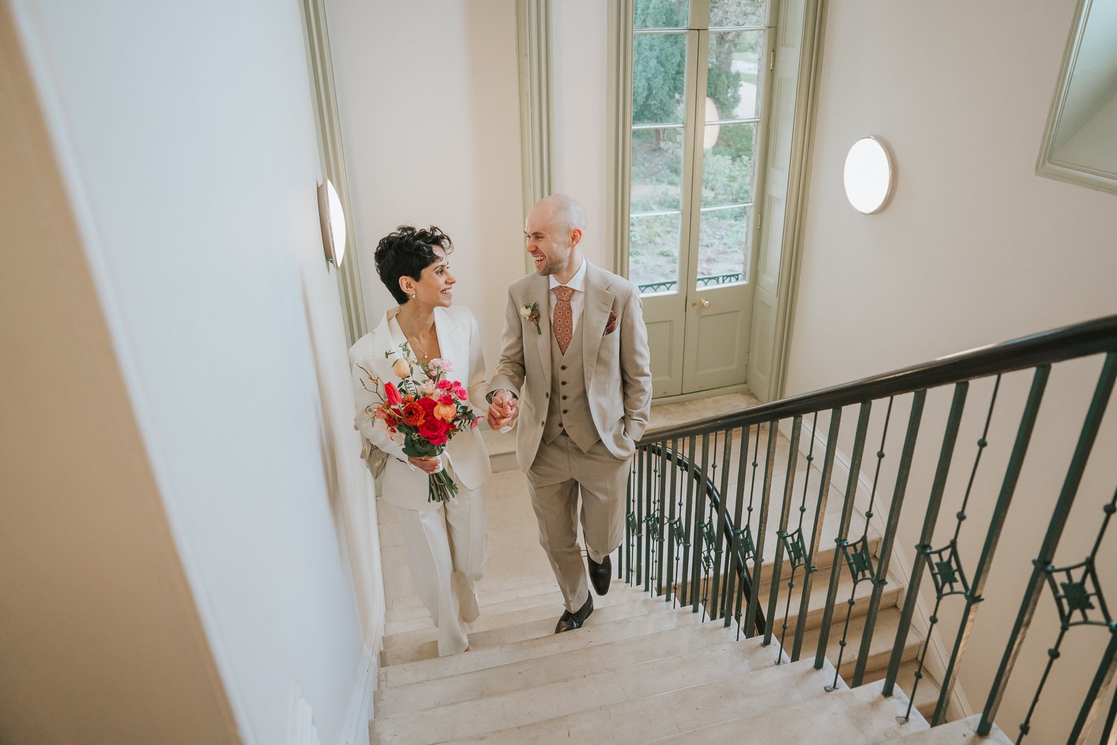  Bride and groom walking hand in hand on Brockwell Hall staircase, soft natural light from tall windows at Brockwell Park wedding venue. 
