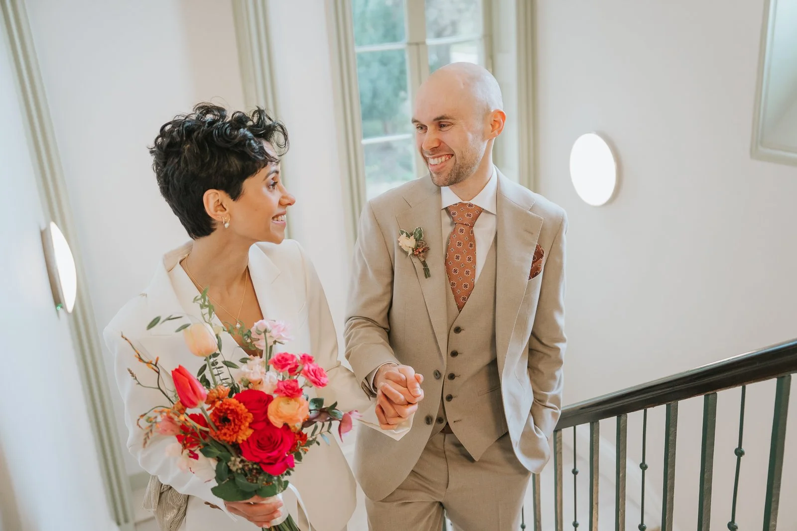  Bride and groom walking hand in hand on Brockwell Hall staircase, soft natural light from tall windows at Brockwell Park wedding venue. 