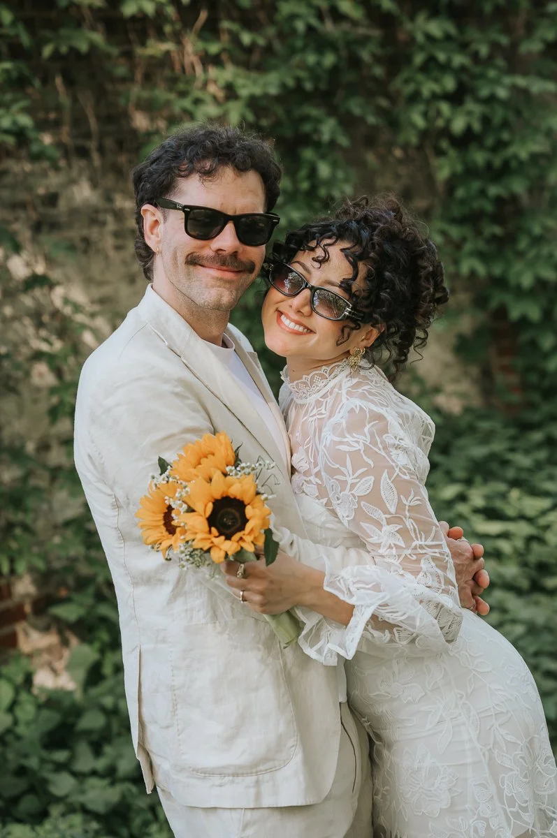  Newly married couple wearing sunglasses holding sunflower bouquet at Tower Hamlets Register Office garden. 