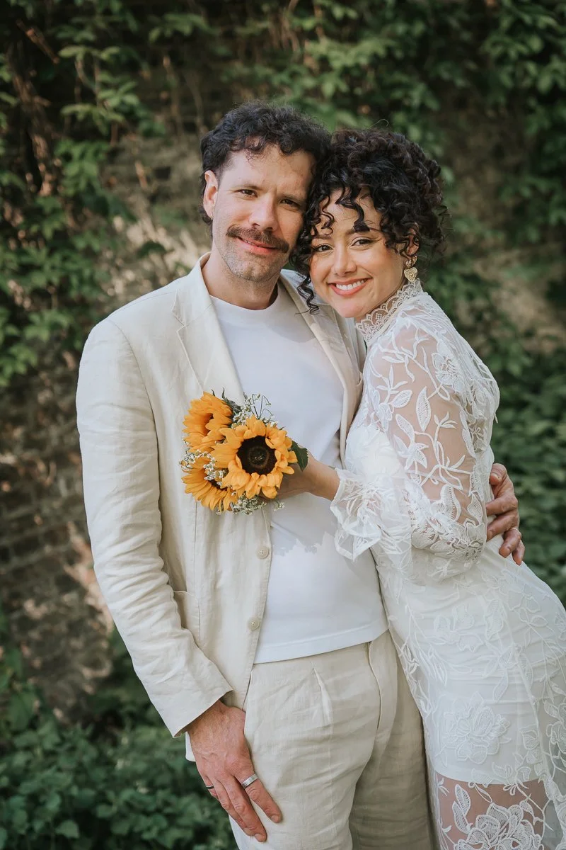  Close-up of bride and groom smiling with sunflower bouquet in outdoor garden at Tower Hamlets Register Office London. 