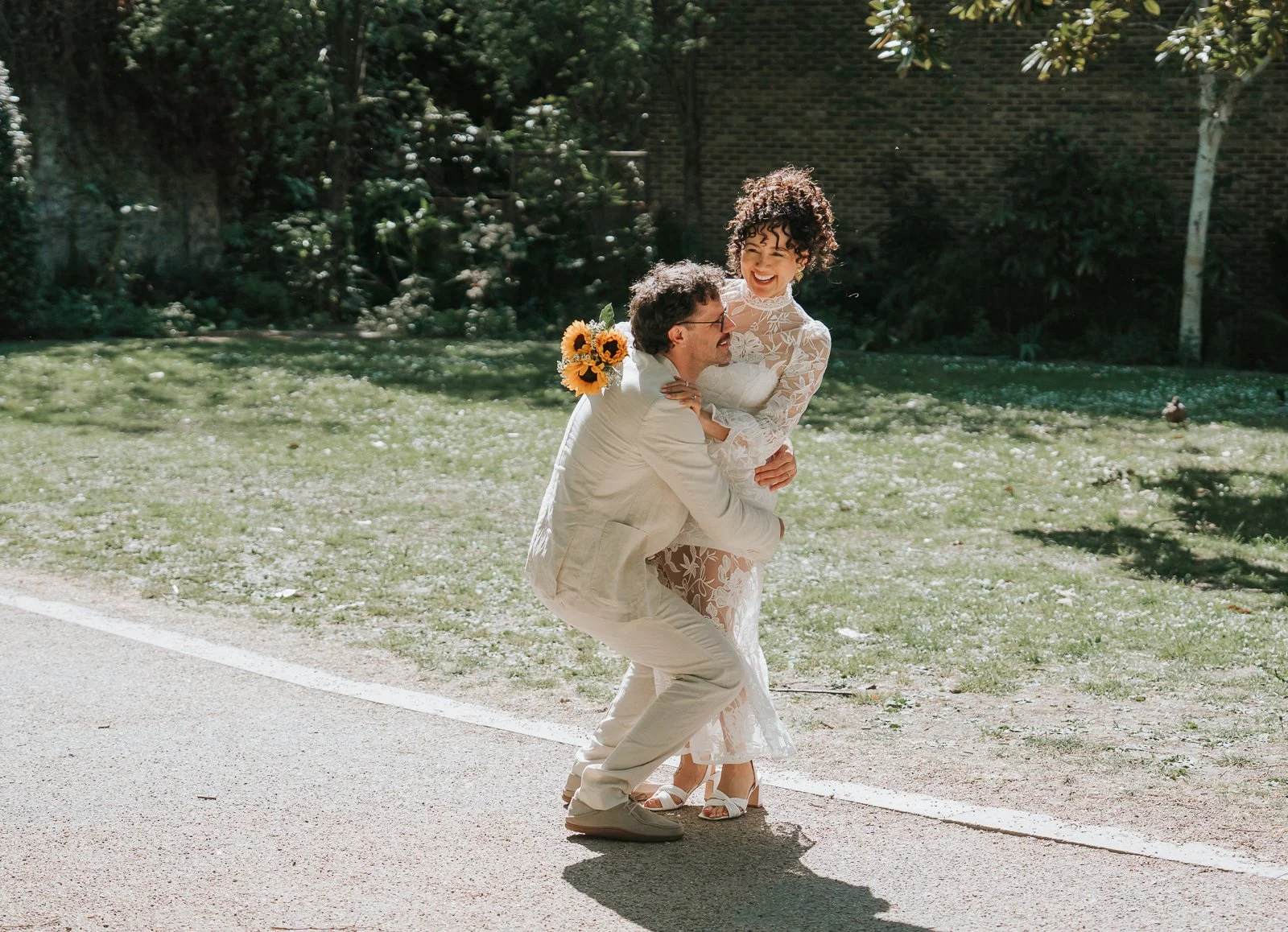  Couple laughing and embracing in sunlit outdoor space at Tower Hamlets Registry Office after wedding ceremony. 