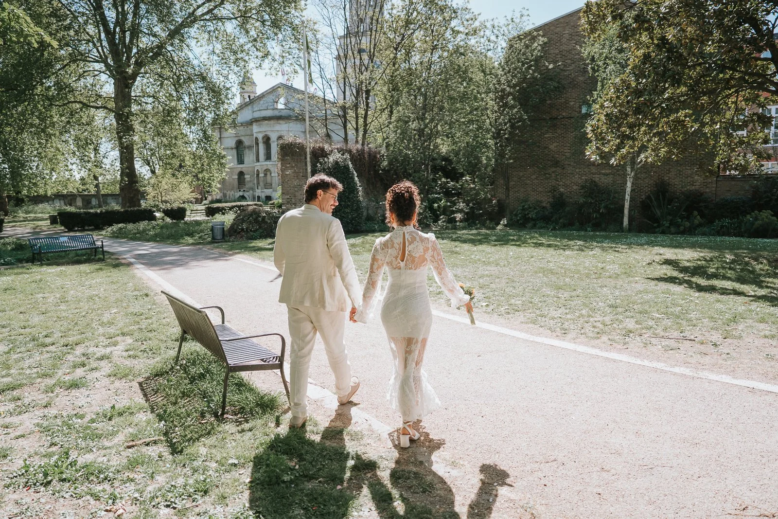  Newly married couple walking hand in hand along leafy path near Tower Hamlets Registry Office East London. 