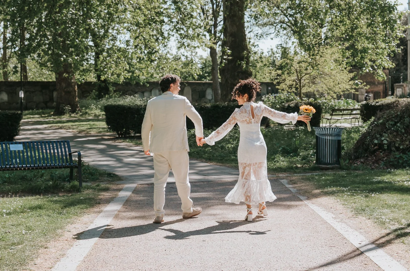  Newly married couple walking hand in hand along leafy path near Tower Hamlets Registry Office East London. 