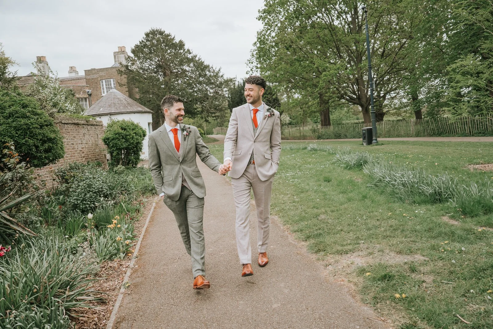  Newly married couple walking hand in hand through the gardens at Morden Park House, surrounded by greenery and spring flowers during a relaxed South London wedding. 