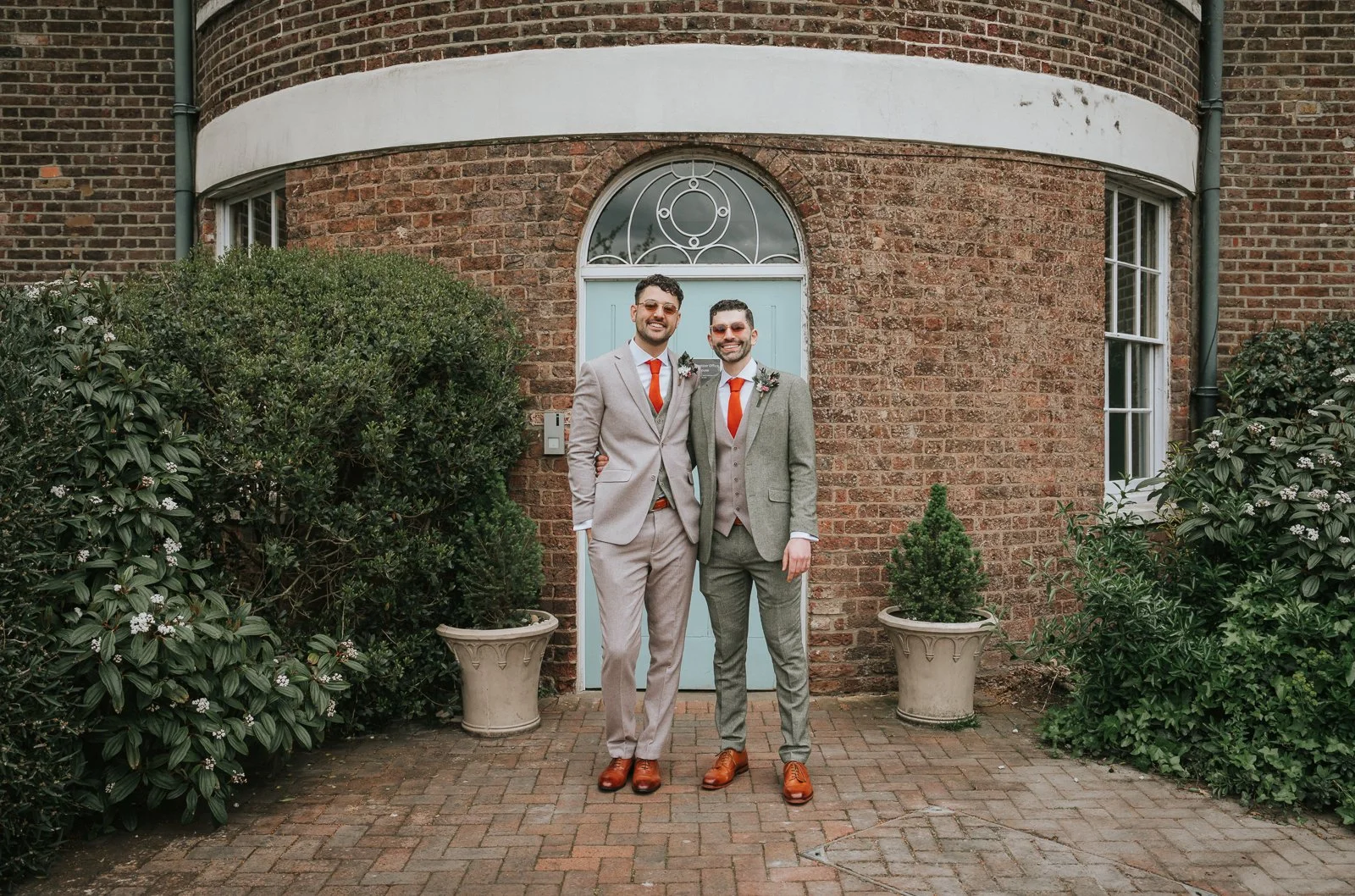  Same-sex couple standing outside Morden Park House after their wedding ceremony, wearing neutral suits with bright orange ties in front of the Georgian manor house entrance. 