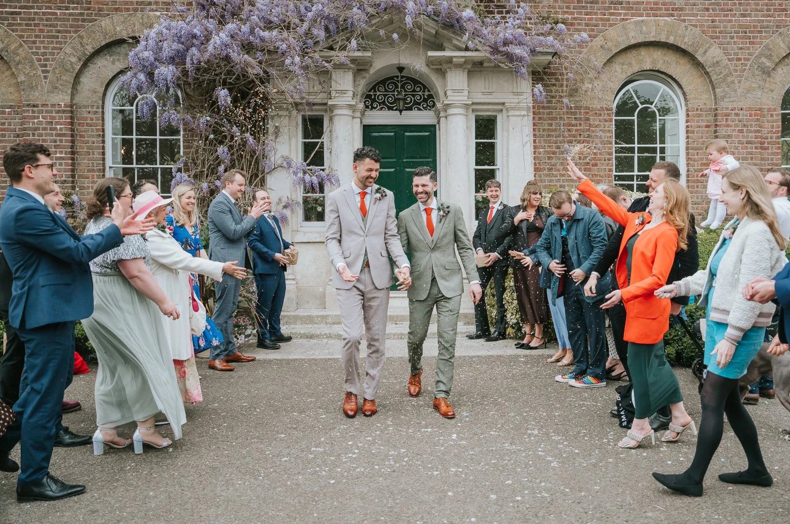  Confetti moment outside Morden Park House as guests celebrate a same-sex wedding beneath blooming wisteria and the Georgian manor house entrance. 