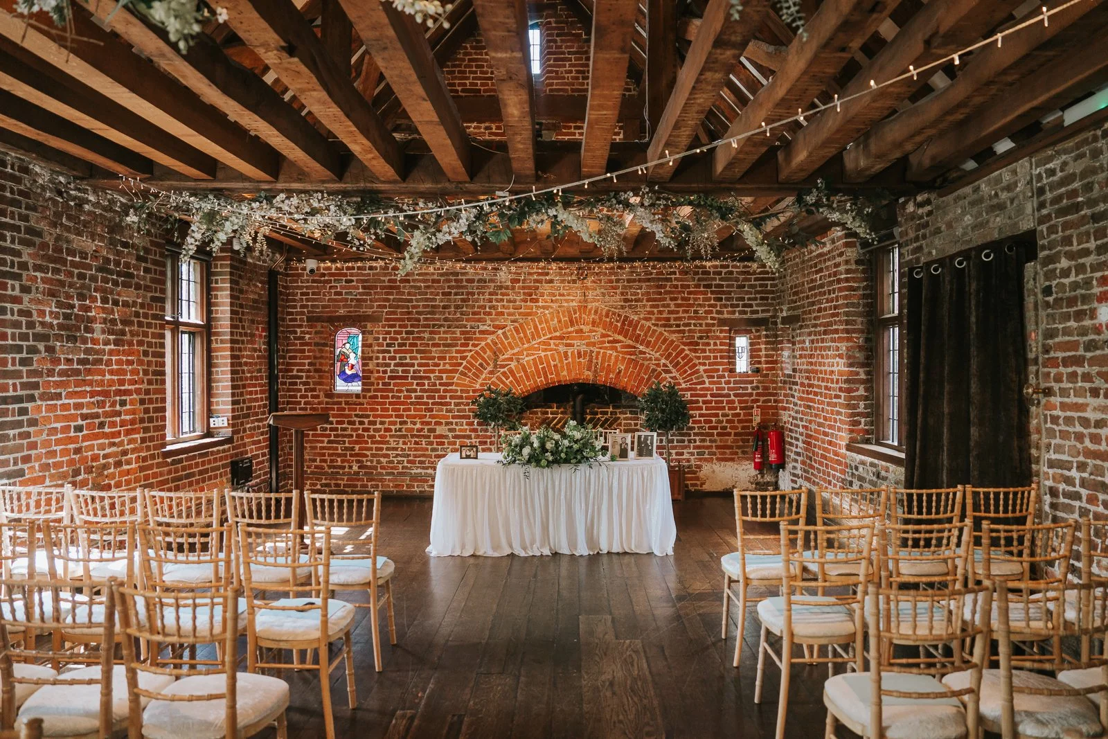  Rustic wedding ceremony setup inside Tudor Barn Eltham with exposed wooden beams, brick walls, floral garlands and candle lights above rows of chairs. 