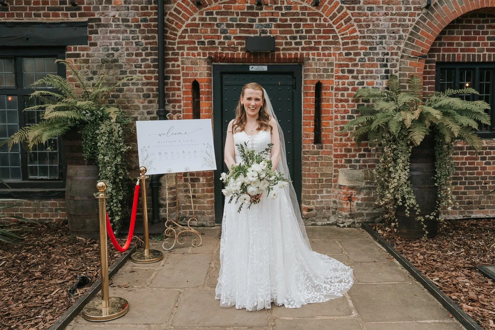  Bride holding a white floral bouquet outside Tudor Barn Eltham with historic brick archways, rustic greenery styling and elegant wedding welcome sign at the barn entrance. 