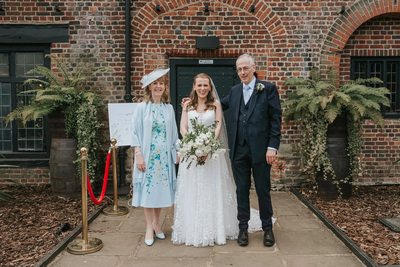  Bride posing with her parents outside Tudor Barn Eltham surrounded by historic brickwork, rustic greenery and elegant wedding styling before the ceremony. 