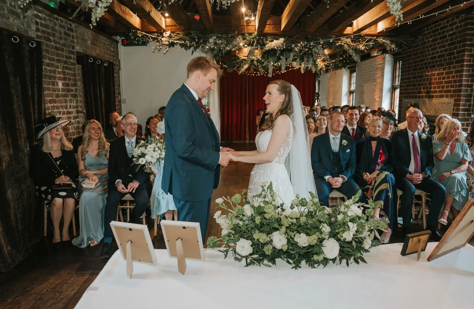  Bride and groom exchanging vows during their wedding ceremony inside Tudor Barn Eltham surrounded by guests, romantic floral decor, exposed wooden beams and warm fairy lights. 