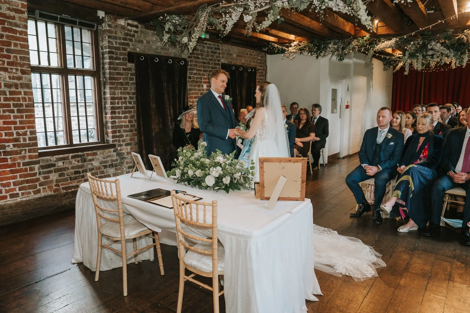  Wedding ceremony at Tudor Barn Eltham with the bride and groom holding hands beneath floral greenery installations, exposed timber beams and rustic brick interiors surrounded by family and friends. 