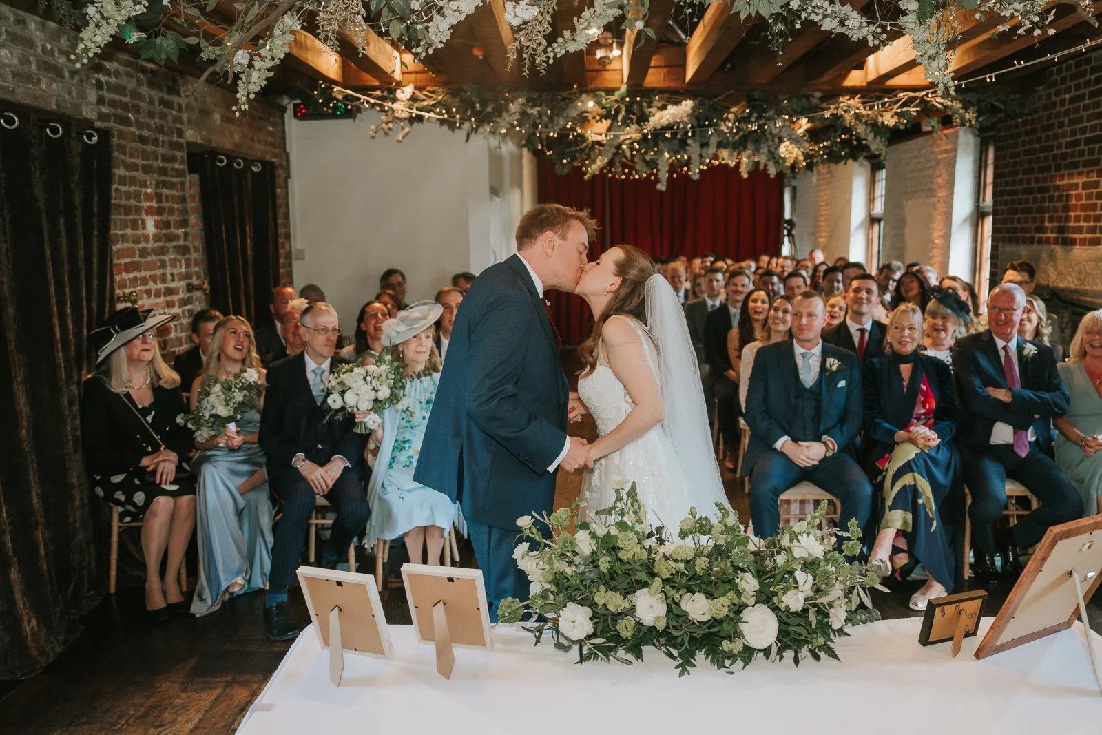  Bride and groom sharing their first kiss during a romantic wedding ceremony at Tudor Barn Eltham surrounded by guests, fairy lights, hanging florals and rustic Tudor barn interiors. 